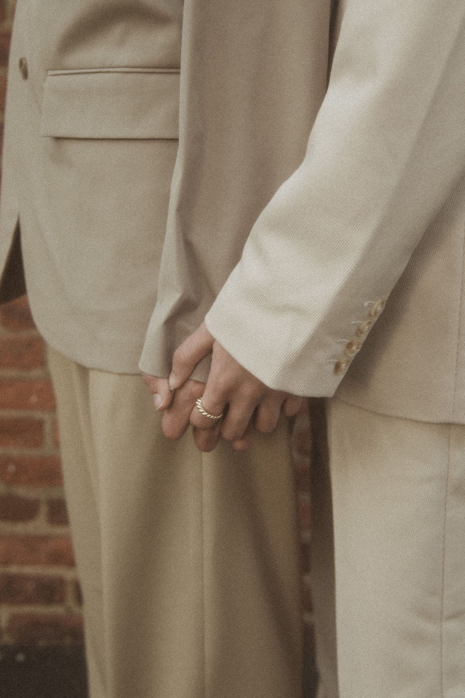 Men in matching neutral colored suits stand in a line showing only their torsos and legs with a brick wall behind them.