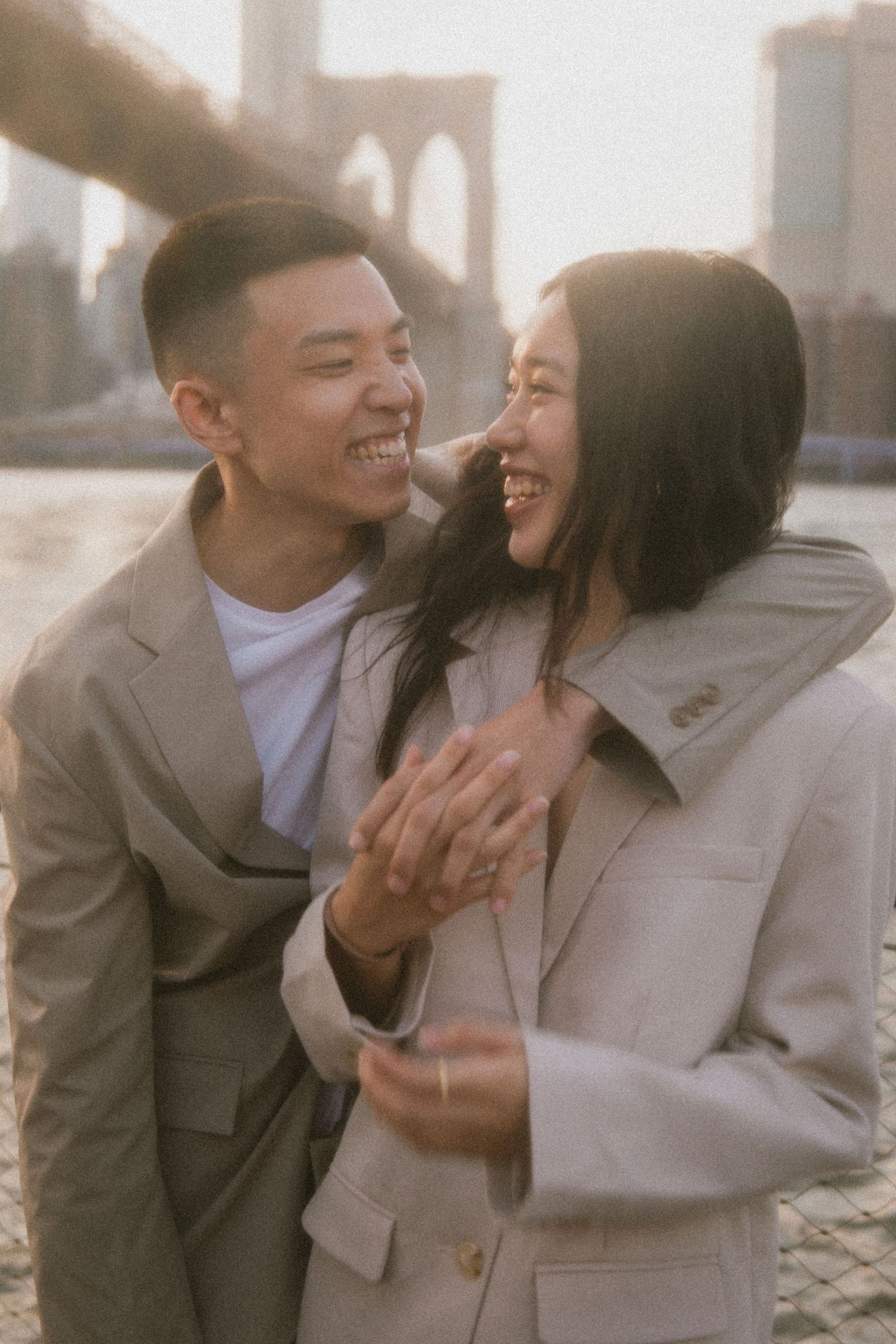 A happy couple laughs together along the waterfront near the Brooklyn Bridge on a misty afternoon.