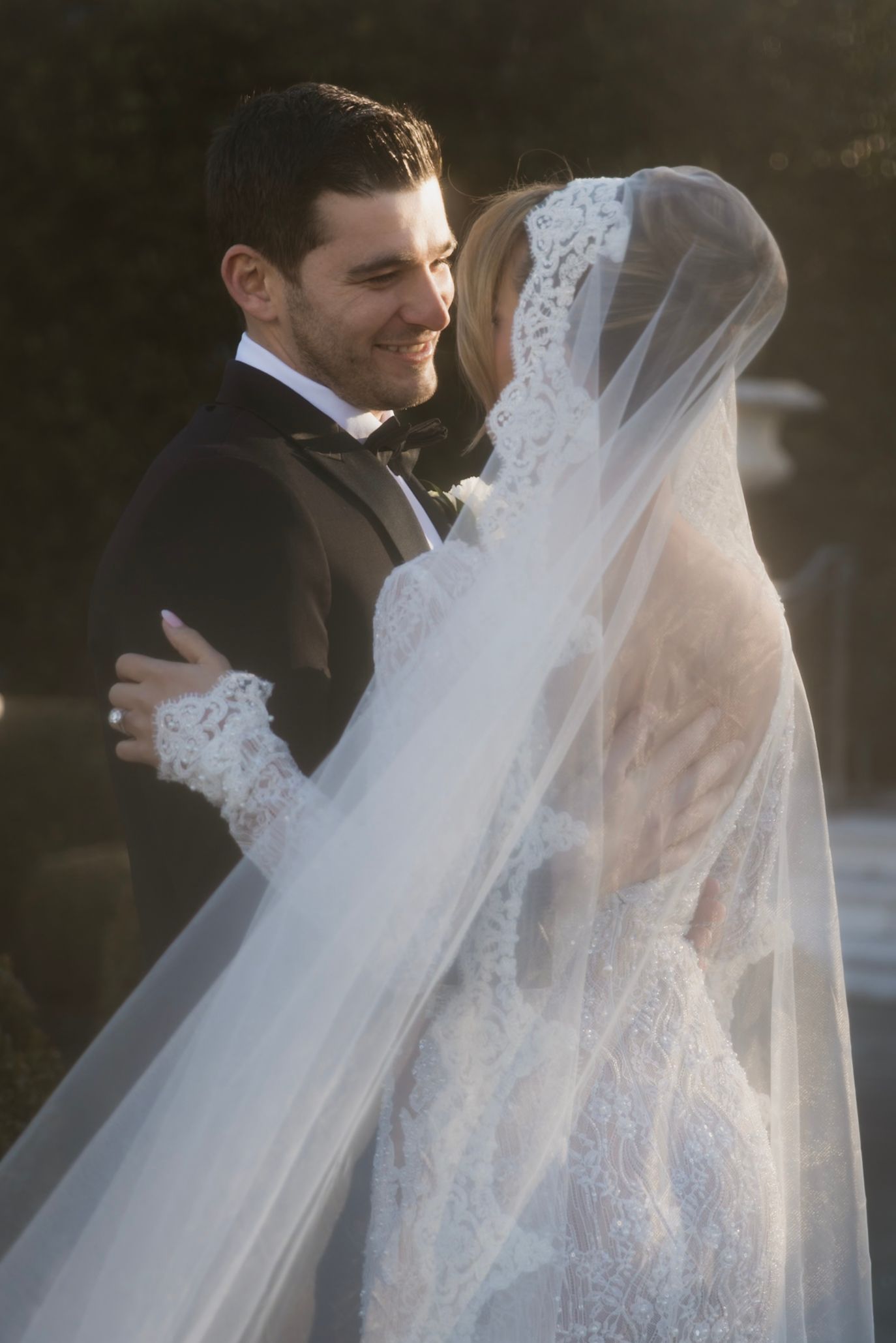 A touching wedding moment showing the bride in a delicate lace veil embracing her groom outdoors at sunset.