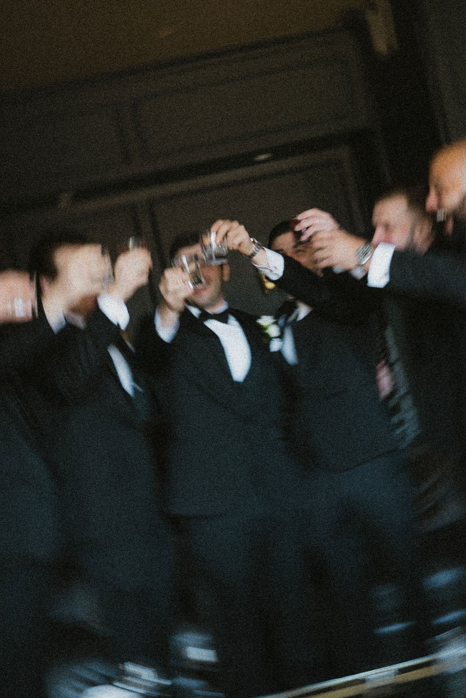 A group in black tuxedos and bow ties taking shots together in a dark, blurred celebration setting.