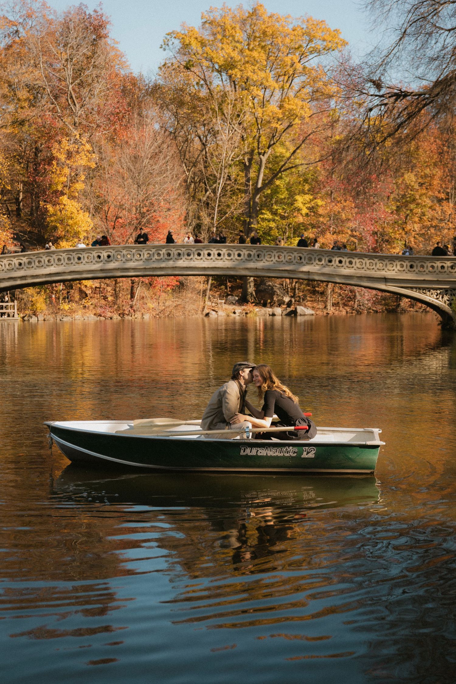 A couple rowing in a green boat on a lake surrounded by trees with vibrant autumn foliage and a stone bridge visible.