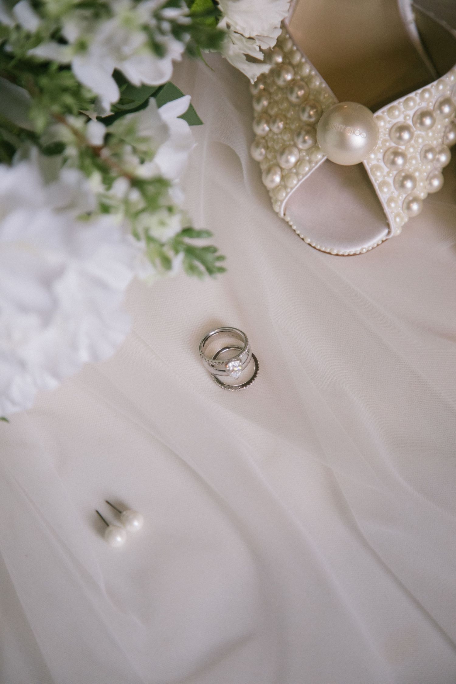 Elegant wedding ring detail shots arranged with delicate pearls, lace, and greenery on light fabric backdrop.