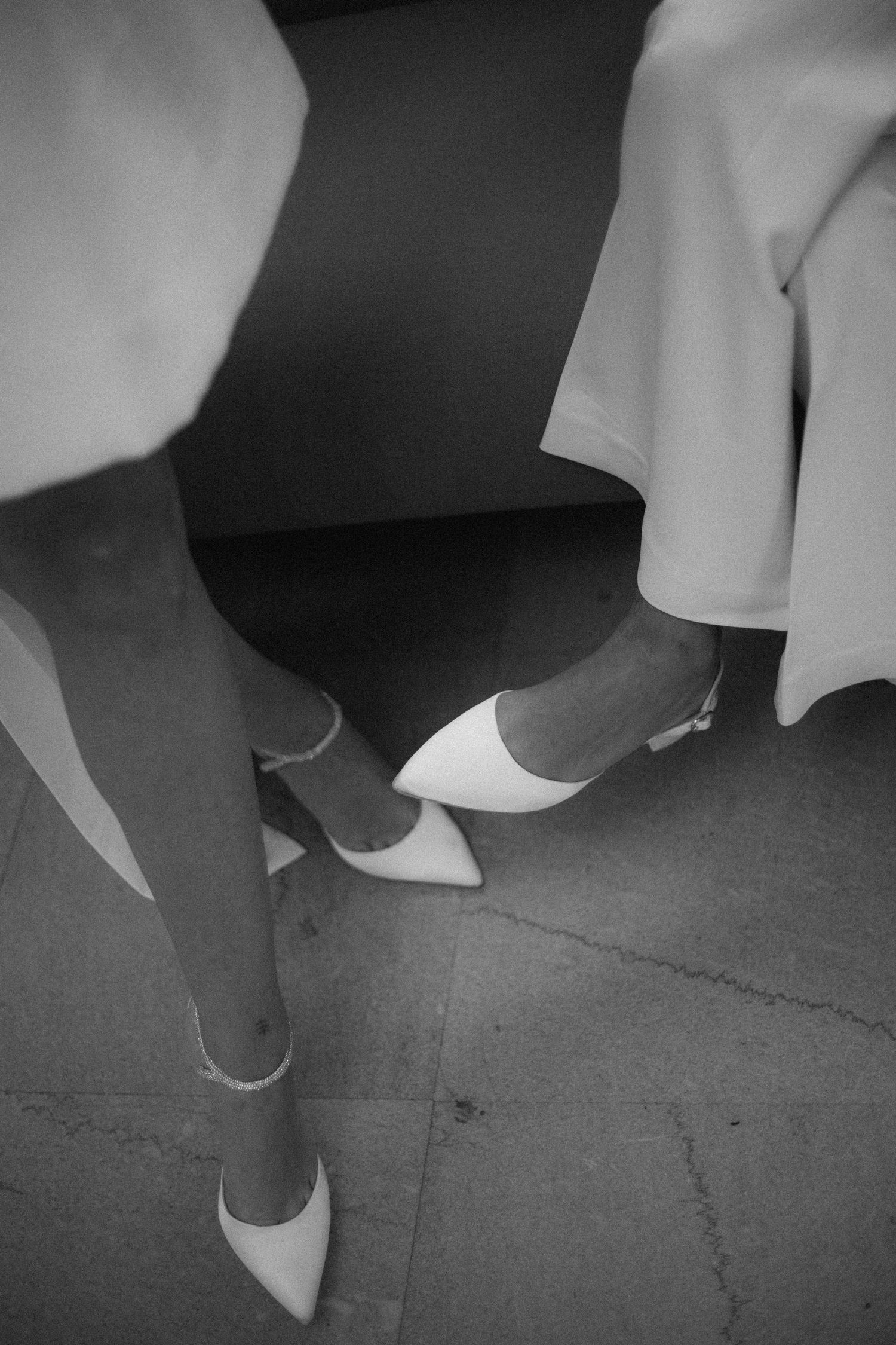Black and white photo showing elegant white shoes and dress hems on a concrete floor.