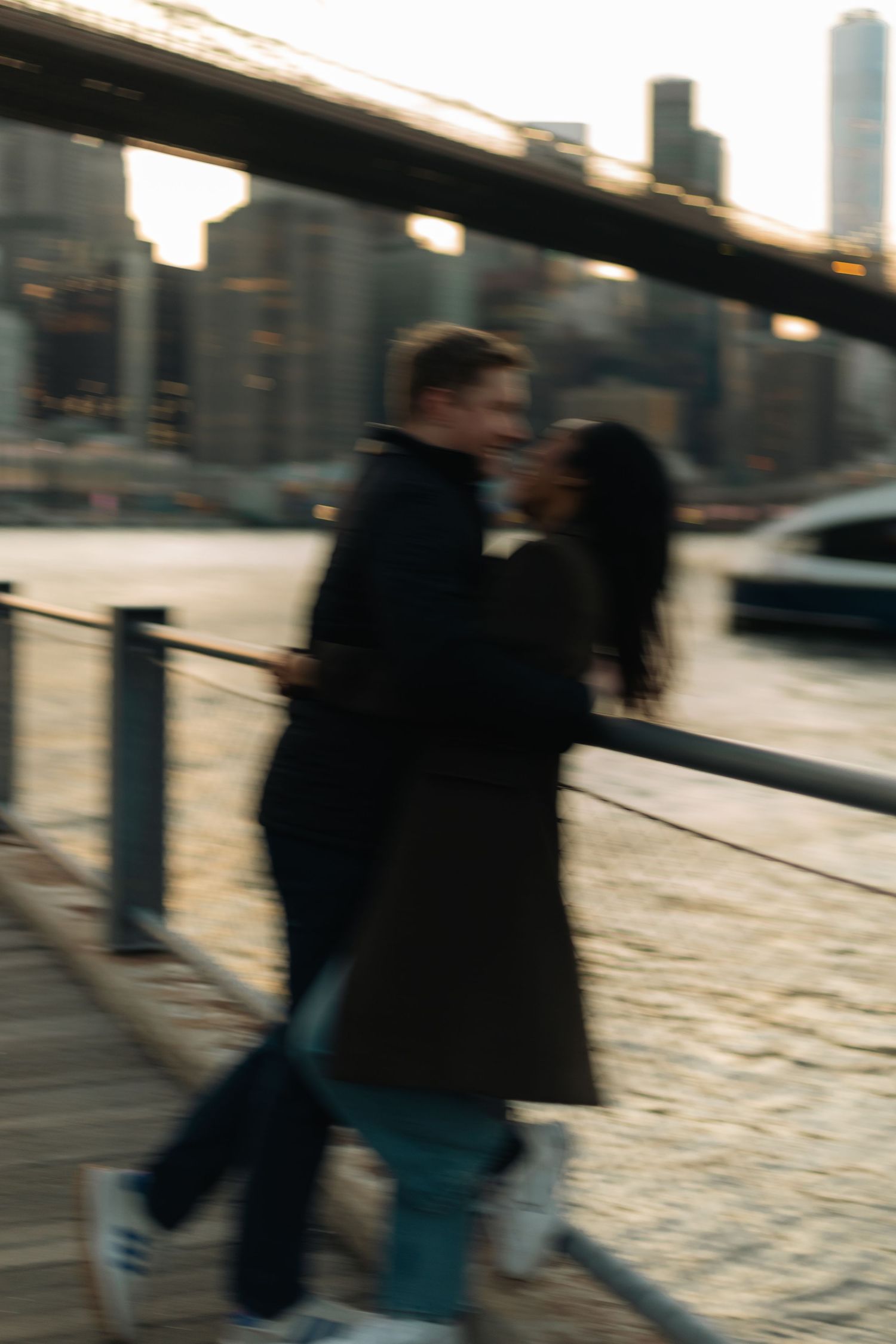 Blurred motion of people walking along an urban waterfront during sunset with city buildings in background.