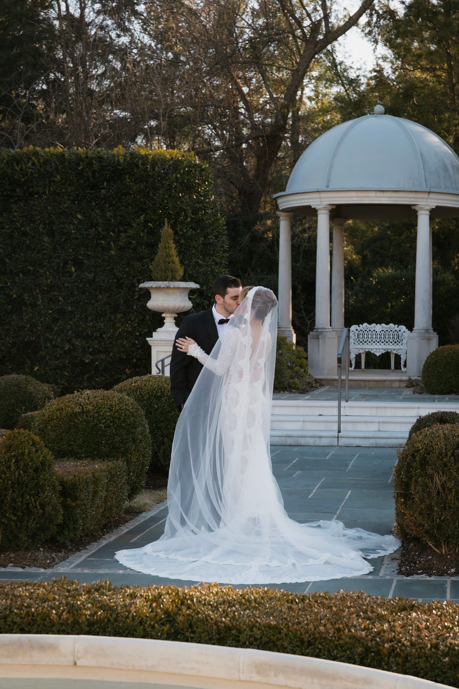 A romantic wedding photoshoot near a white gazebo surrounded by manicured gardens at sunset.