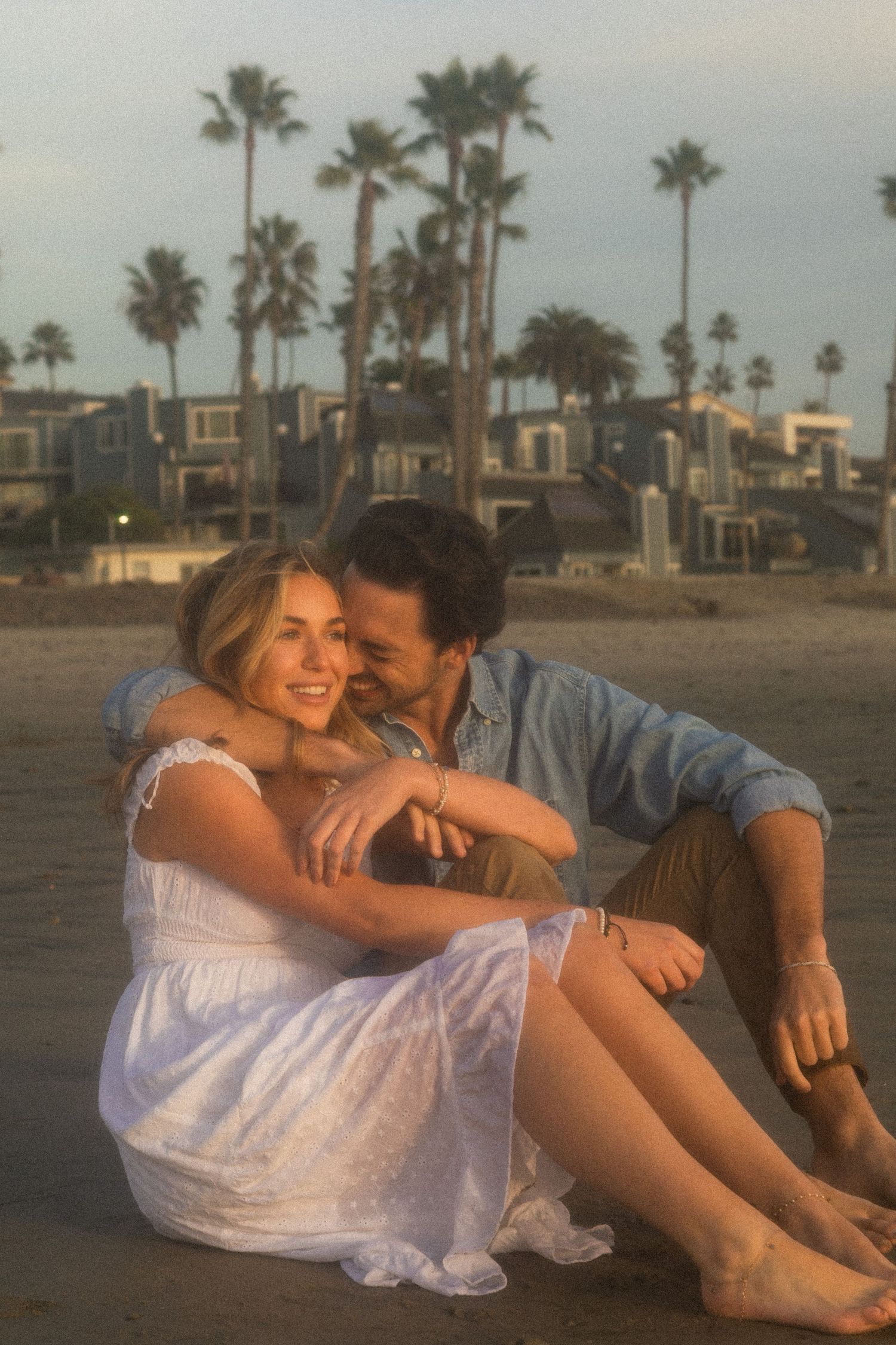 A couple sits close together on a beach at sunset with palm trees and beachfront homes in the background.