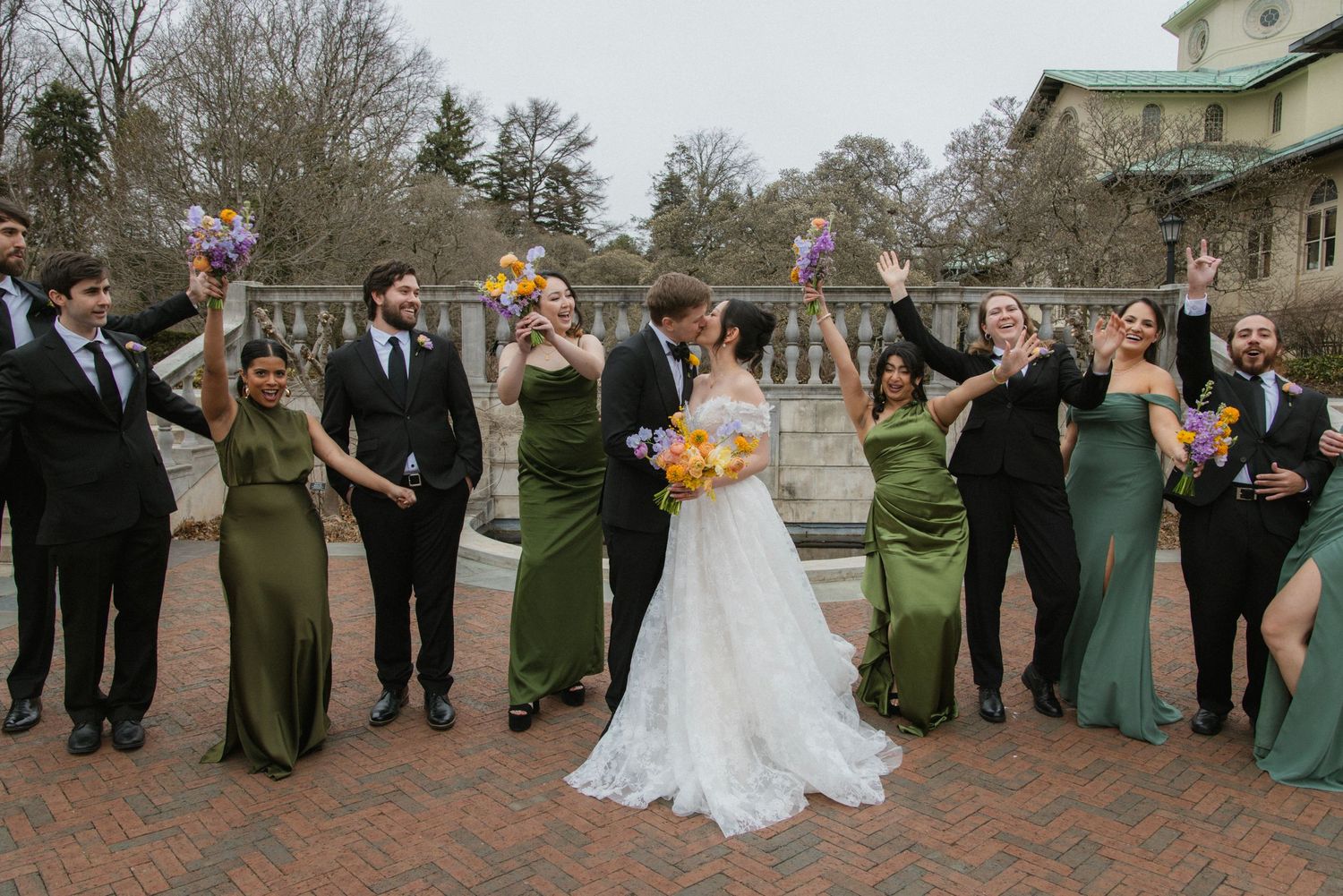 Wedding party poses outdoors with bridesmaids in green dresses and groomsmen in black suits.