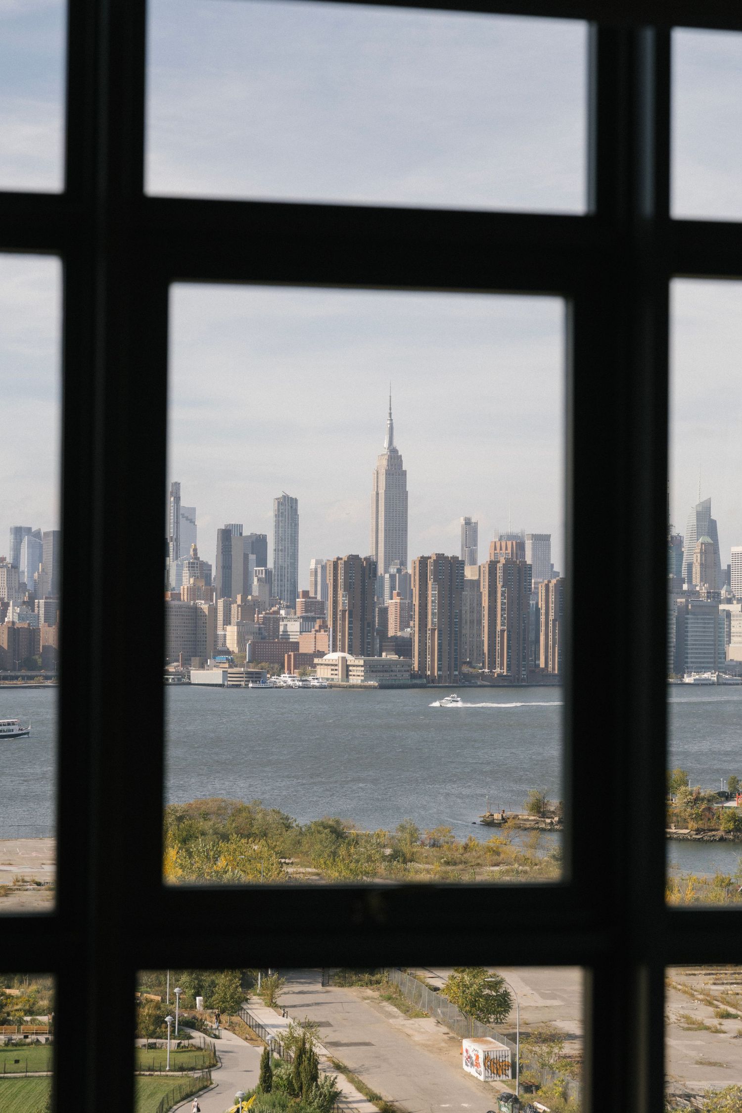 View of New York City skyline through window frame showing Empire State Building and Hudson River on sunny day.