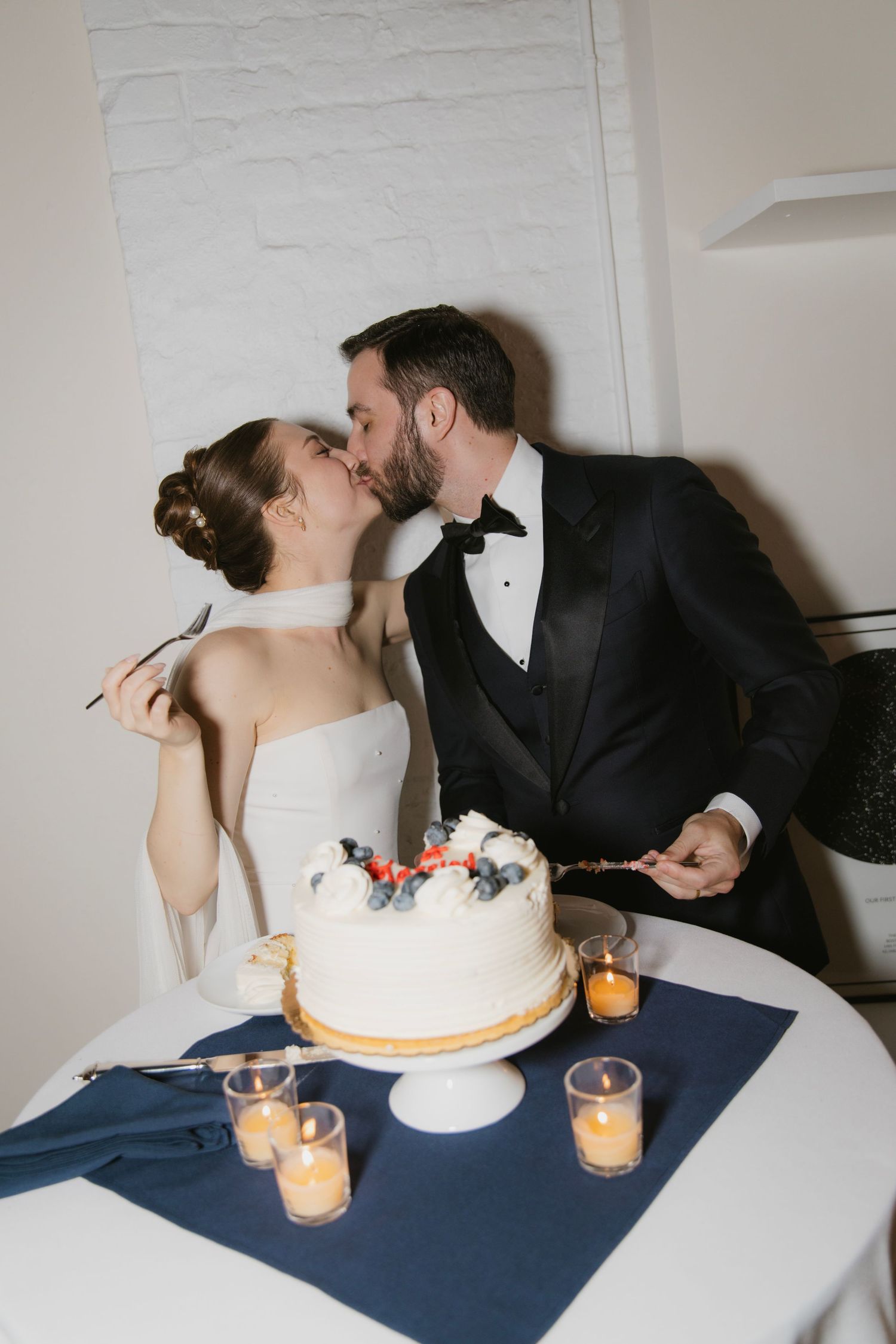 A newlywed couple shares a romantic kiss beside their simple white wedding cake decorated with coral flowers and surrounded by candles.