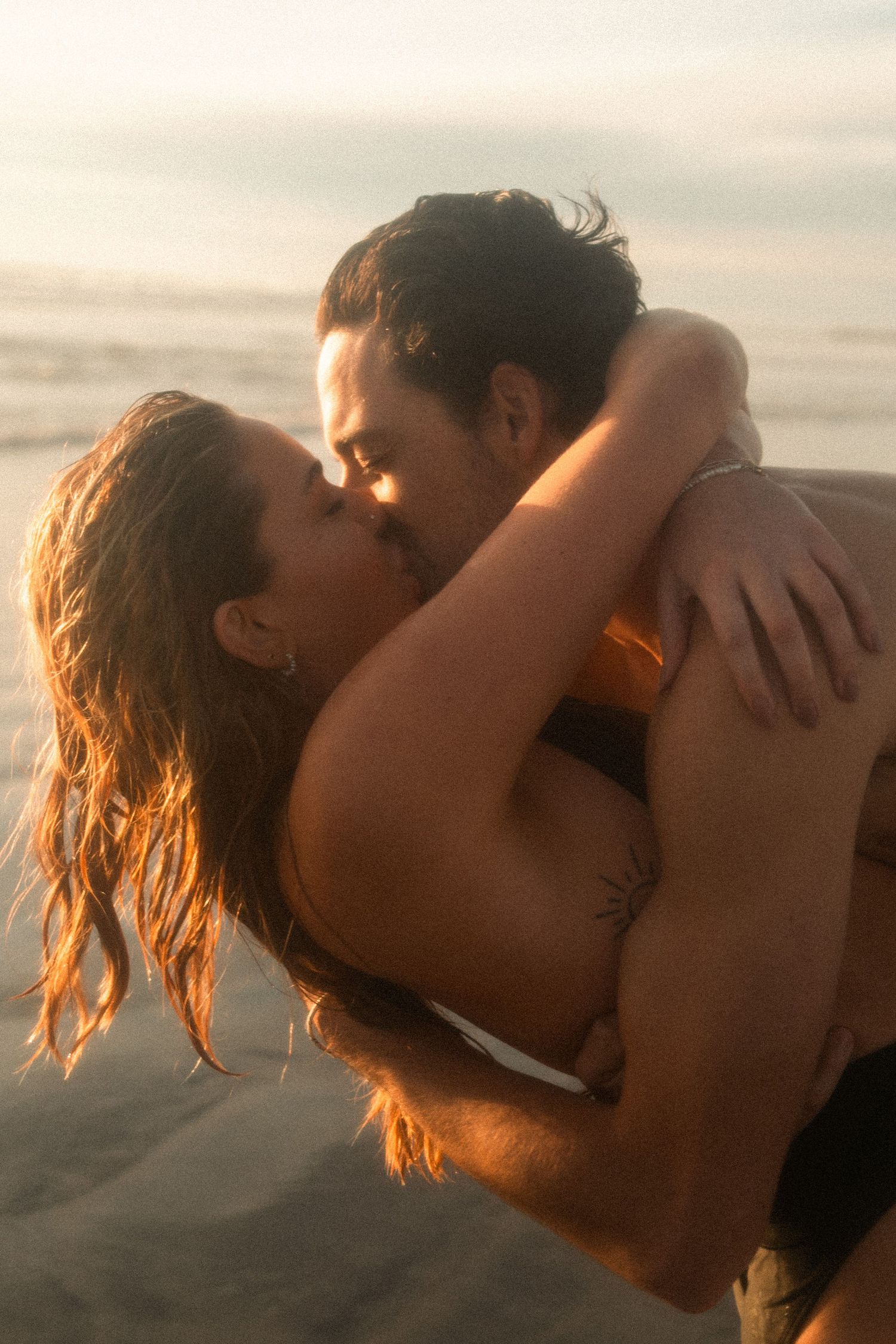 A couple shares a passionate kiss at sunset on the beach with golden light illuminating their silhouettes.
