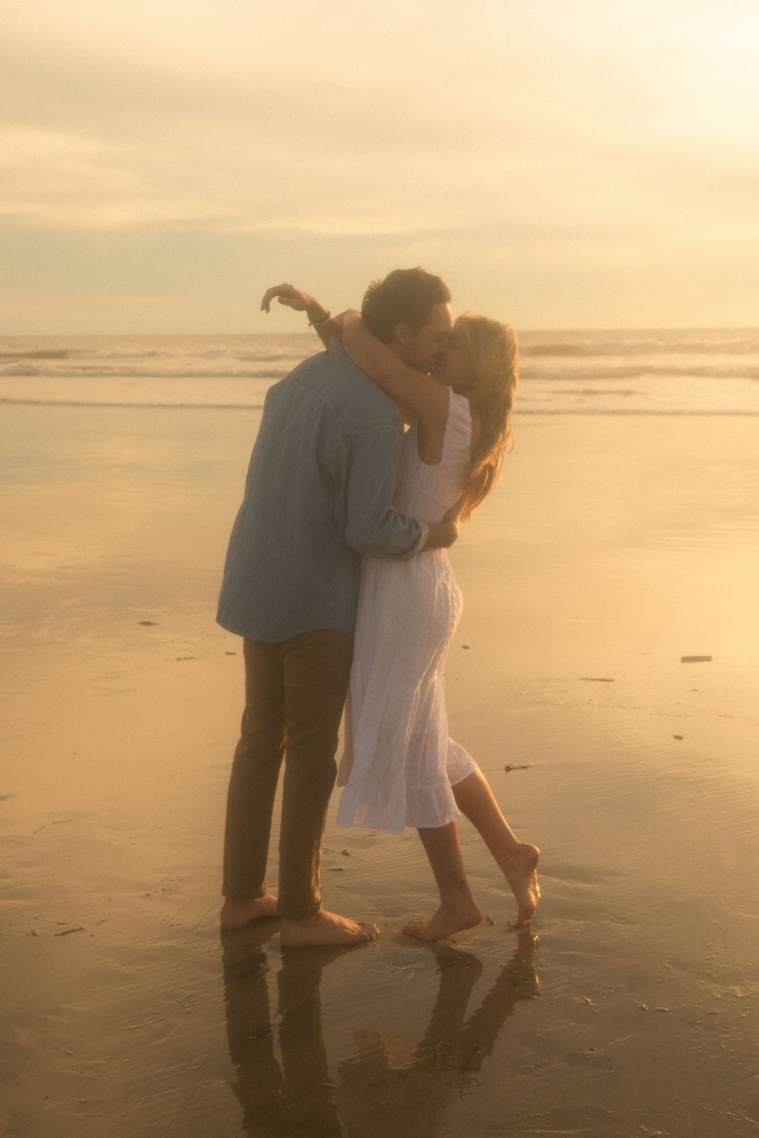Two people walk together on a misty beach during golden hour sunset, looking down at the sand.
