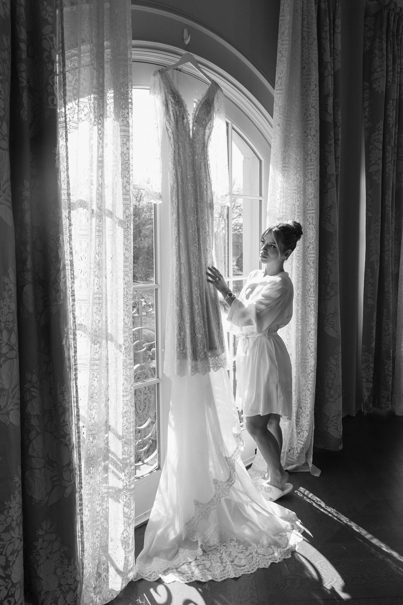 Black and white photo showing a bride in a flowing white wedding gown standing by sunlit curtained windows.