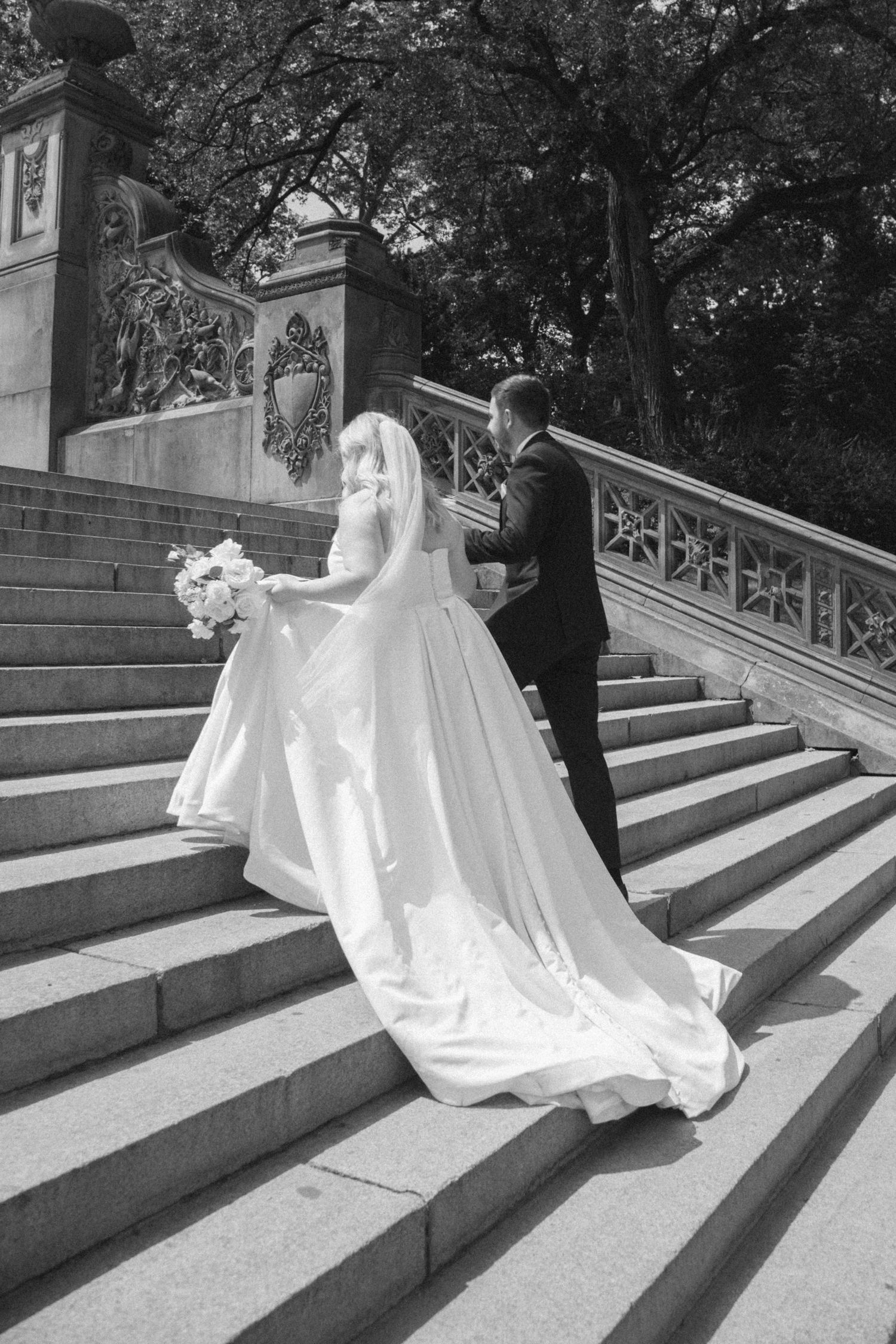 Black and white photo of a couple ascending ornate stone stairs in wedding attire.