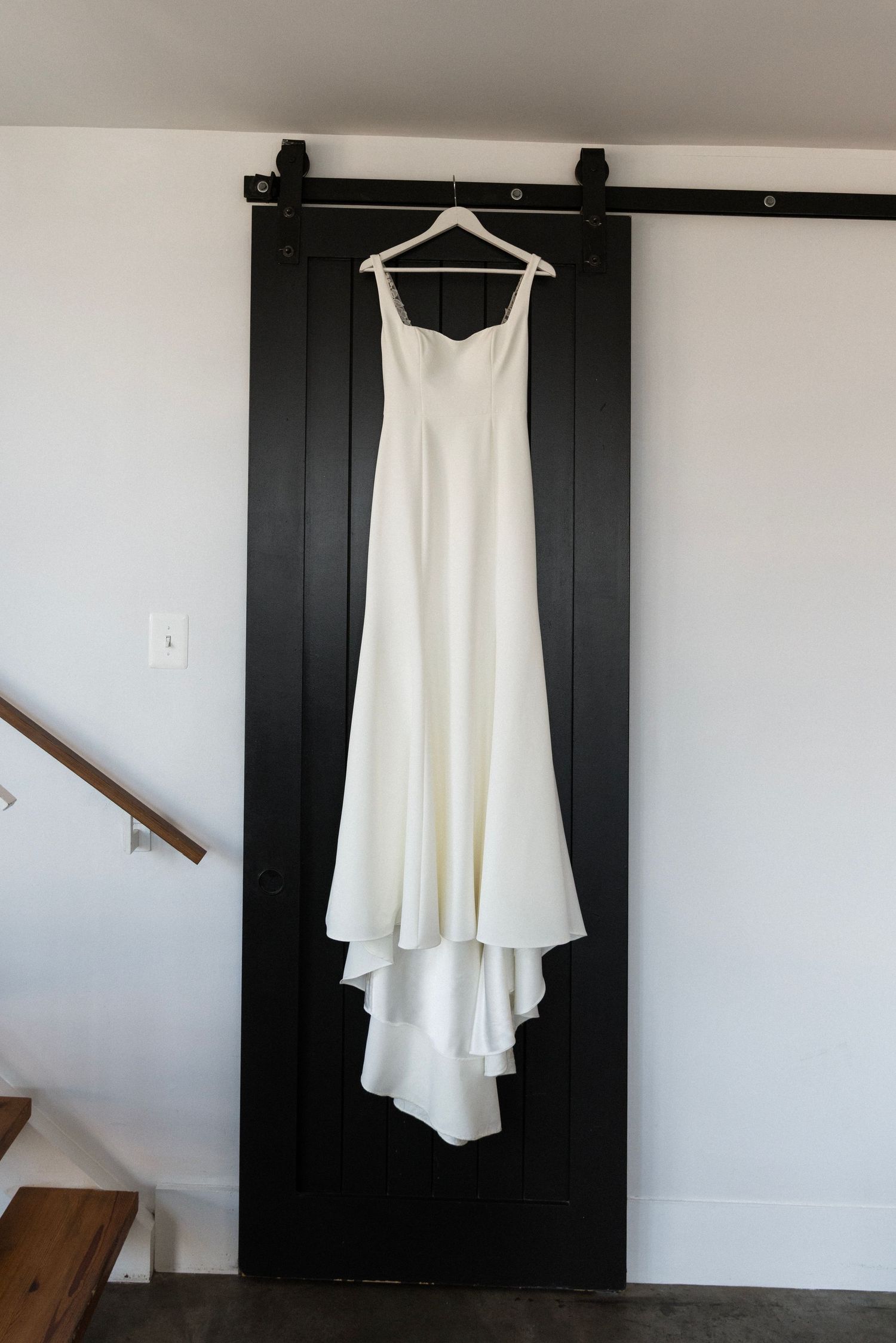 A simple white wedding dress hangs elegantly on a hanger against a black sliding barn door in a minimalist setting.