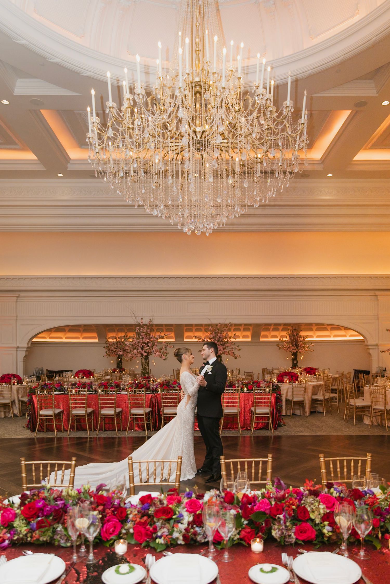 Elegant ballroom wedding reception with crystal chandeliers and vibrant red floral centerpieces.