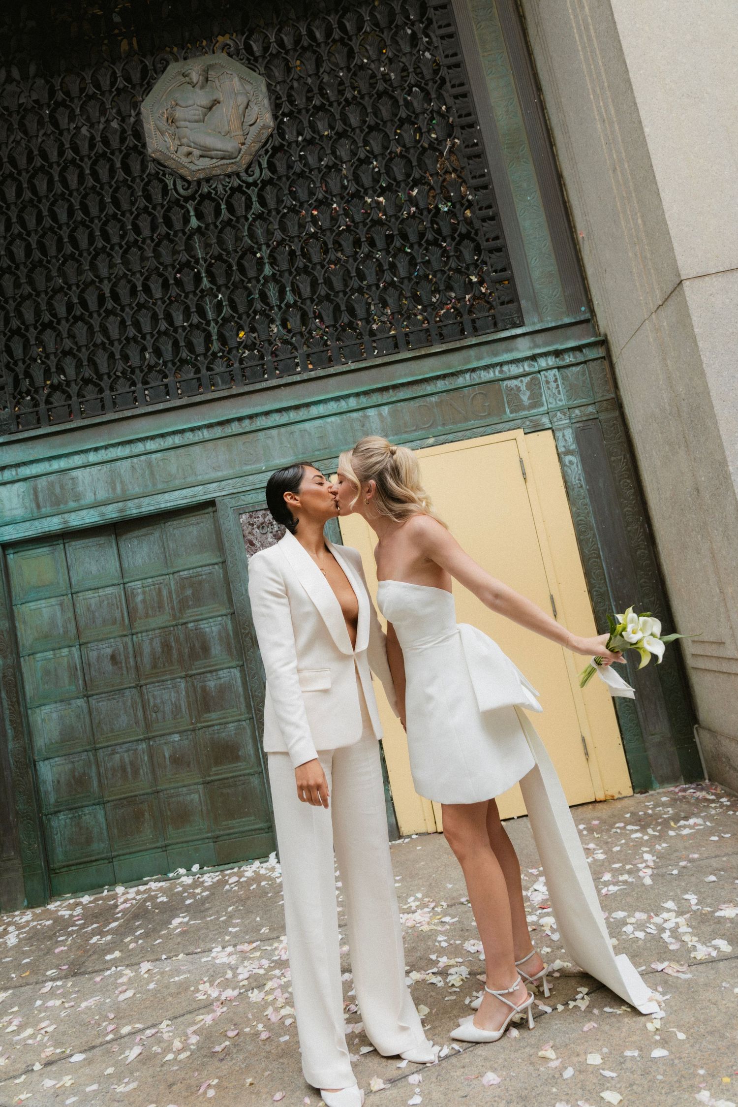 A romantic couple in matching white outfits share a tender moment outside a vintage door with flower petals scattered around.