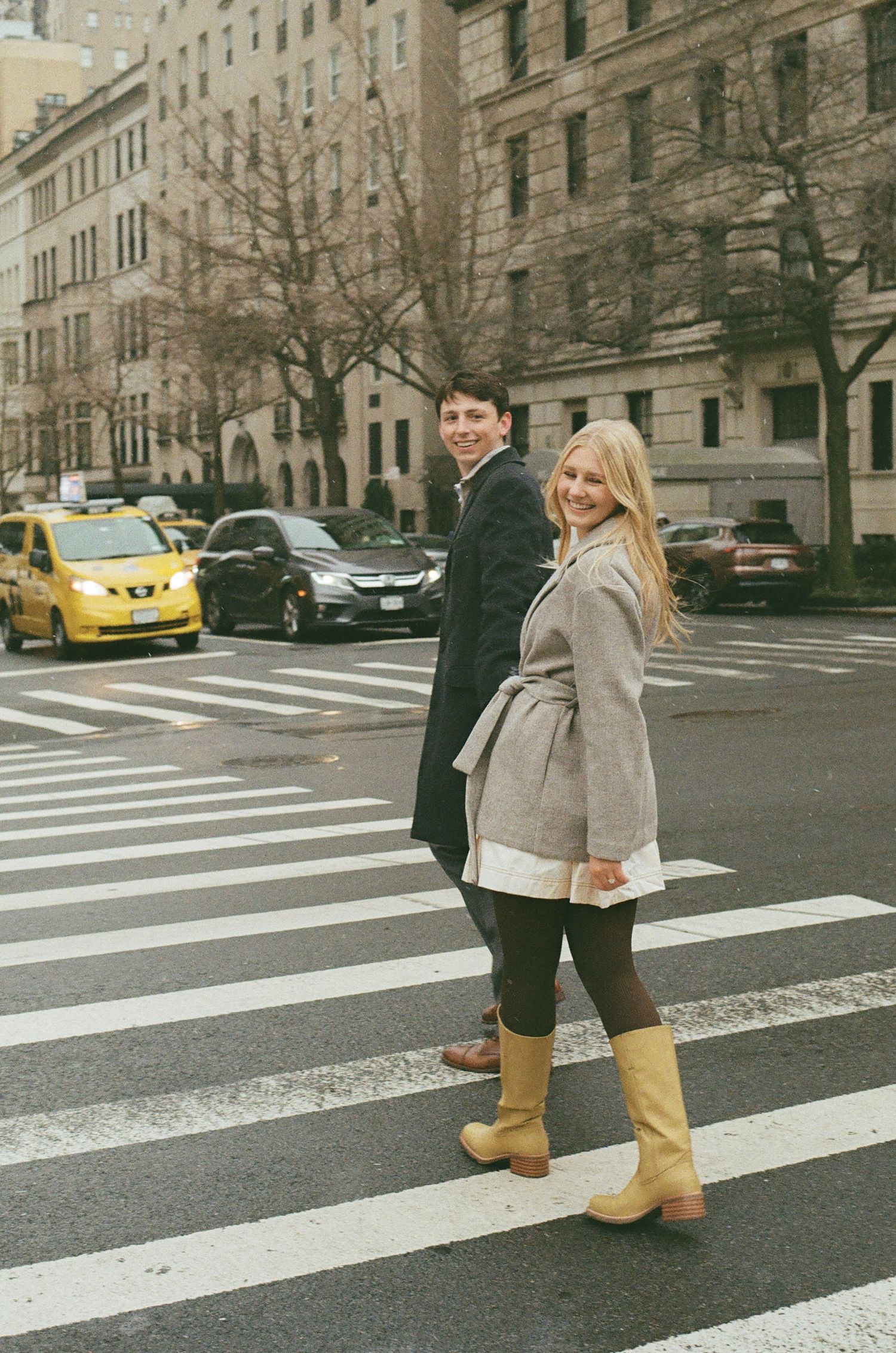 A couple crosses a crosswalk in New York City with a yellow taxi visible in the background.