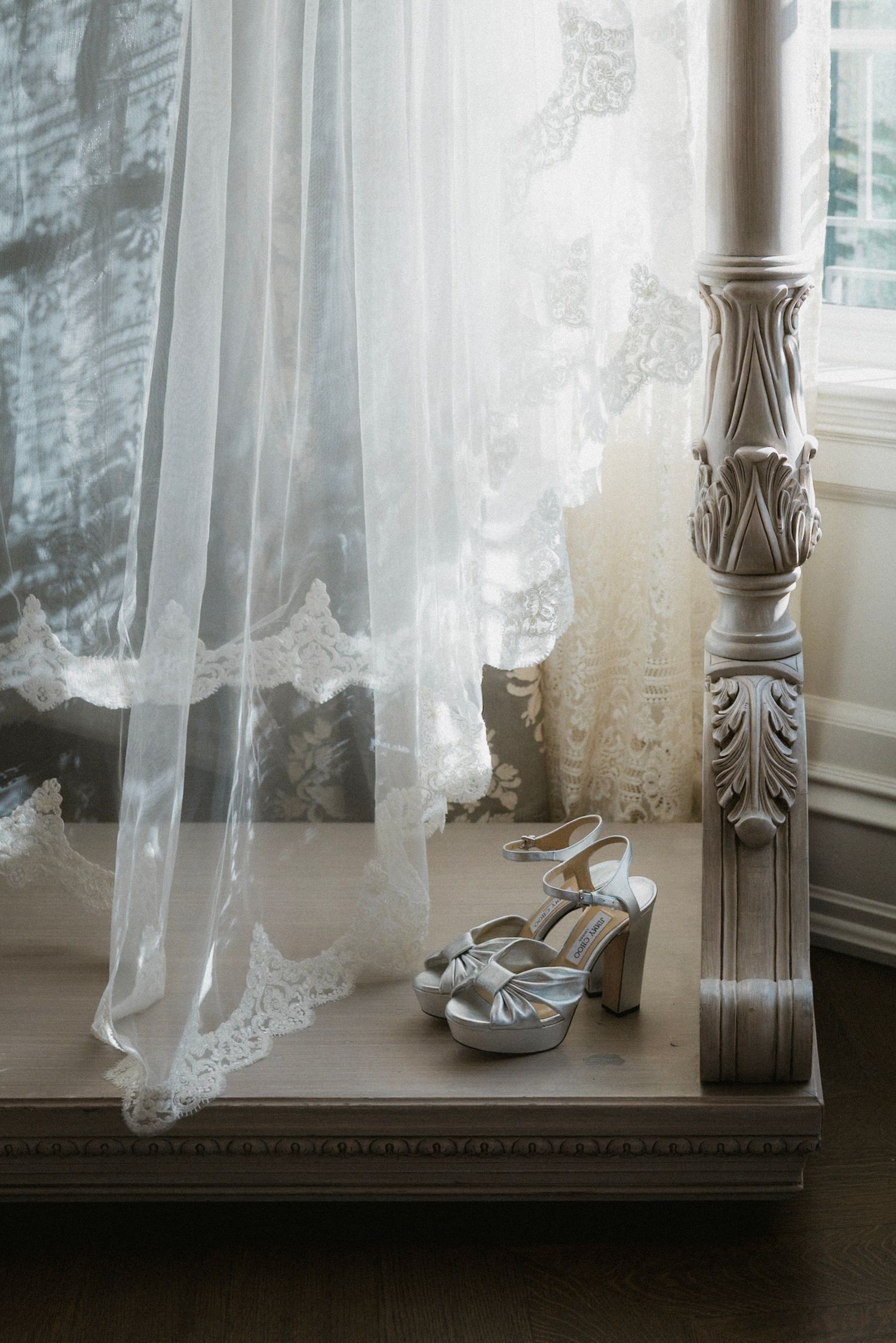 Wedding shoes and lace veil draped near ornate white column by window in natural light.