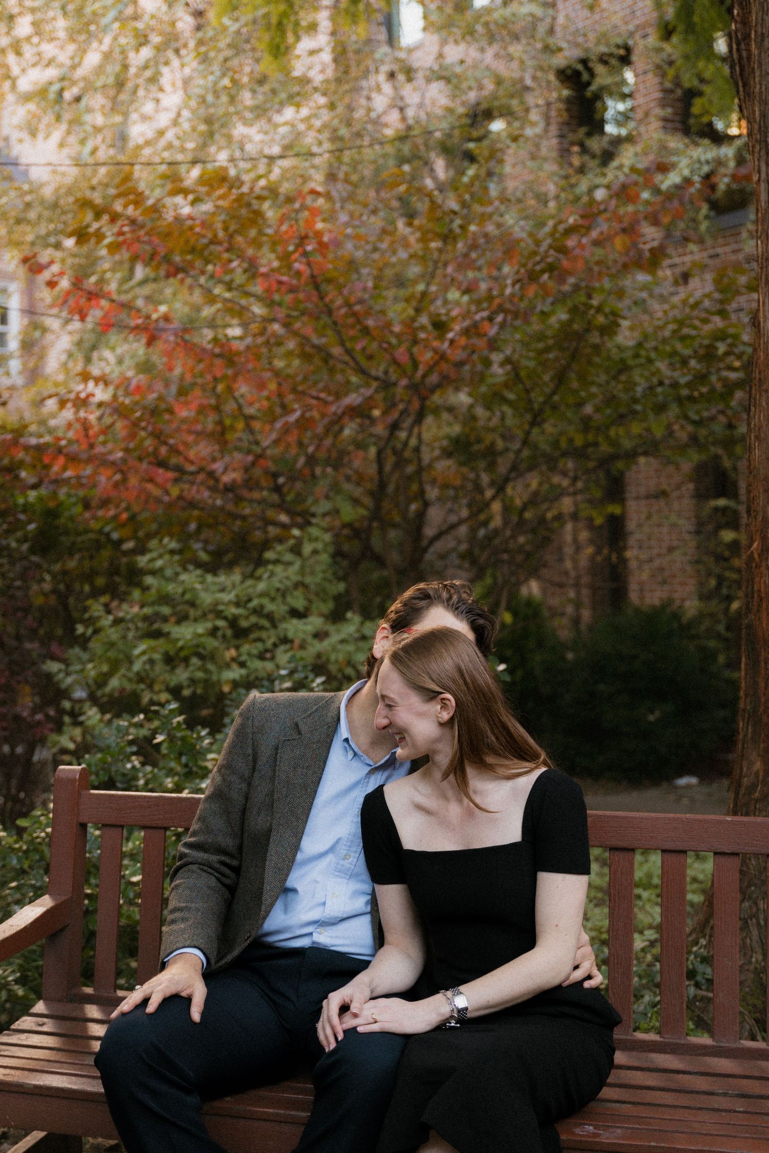 A couple shares an intimate moment while sitting together on a wooden bench in a garden setting with autumn foliage.