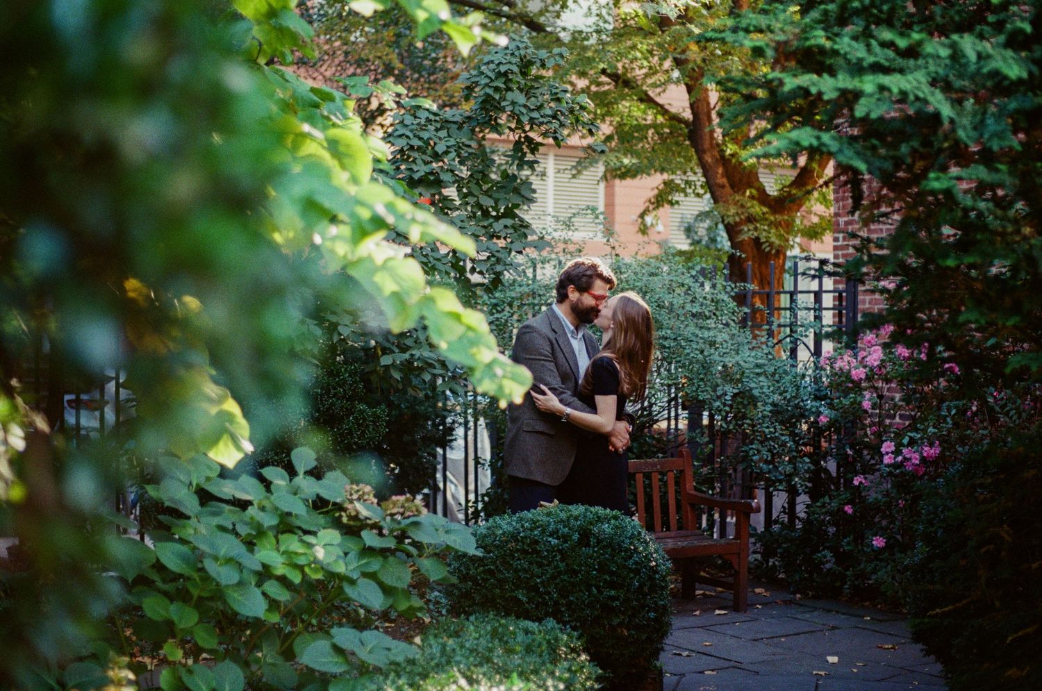 Lush green garden pathway leads to a brick building entrance through overhanging foliage and trees.