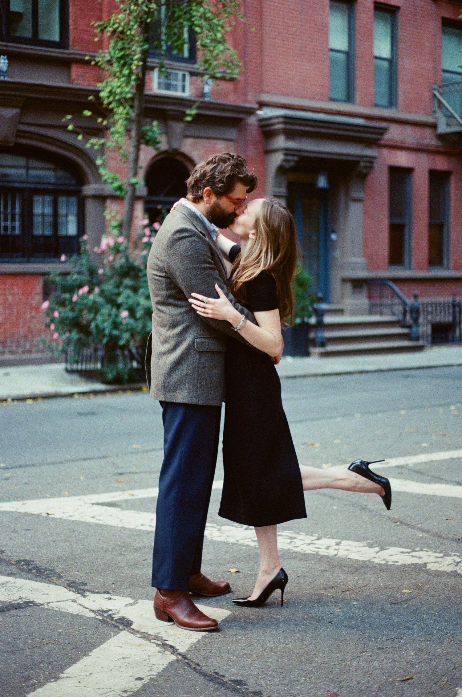 A romantic couple shares a kiss on a city street with brick buildings in the background.