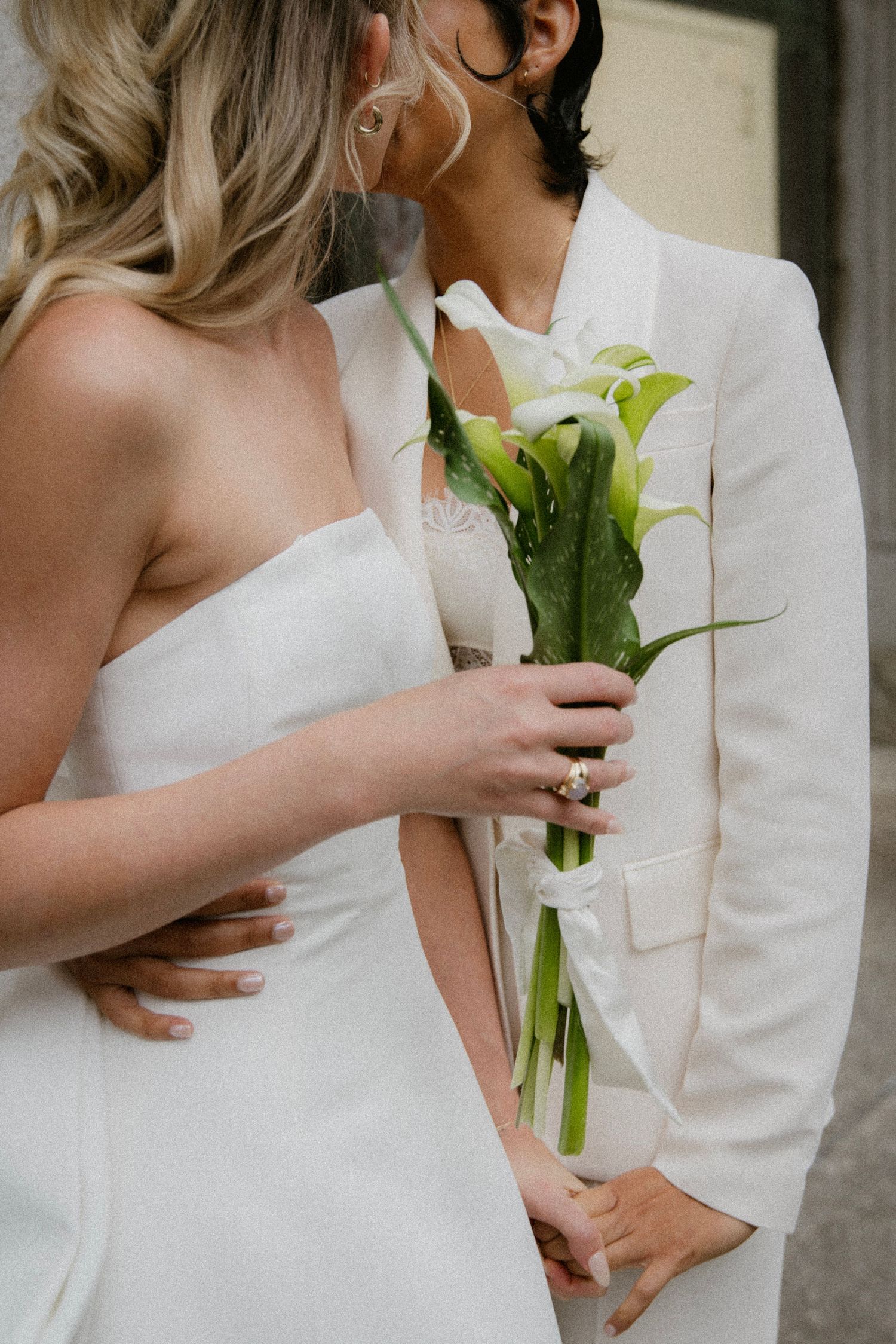 Close-up of newlyweds in white attire embracing while holding elegant calla lily bouquet during intimate wedding moment.