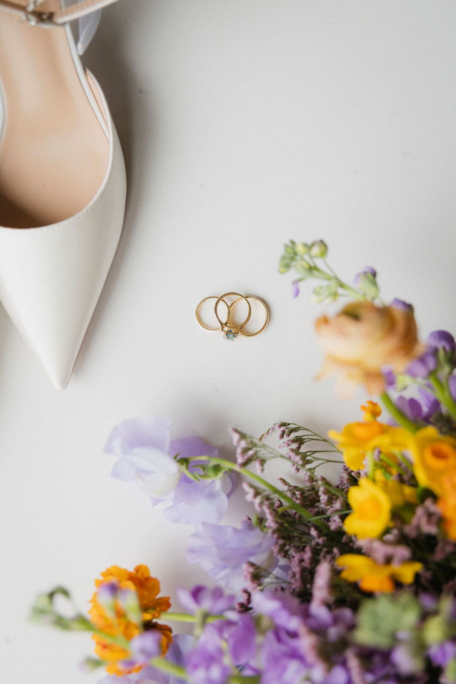 Wedding rings rest beside elegant white bridal shoes with colorful purple, yellow and orange flowers in soft focus.
