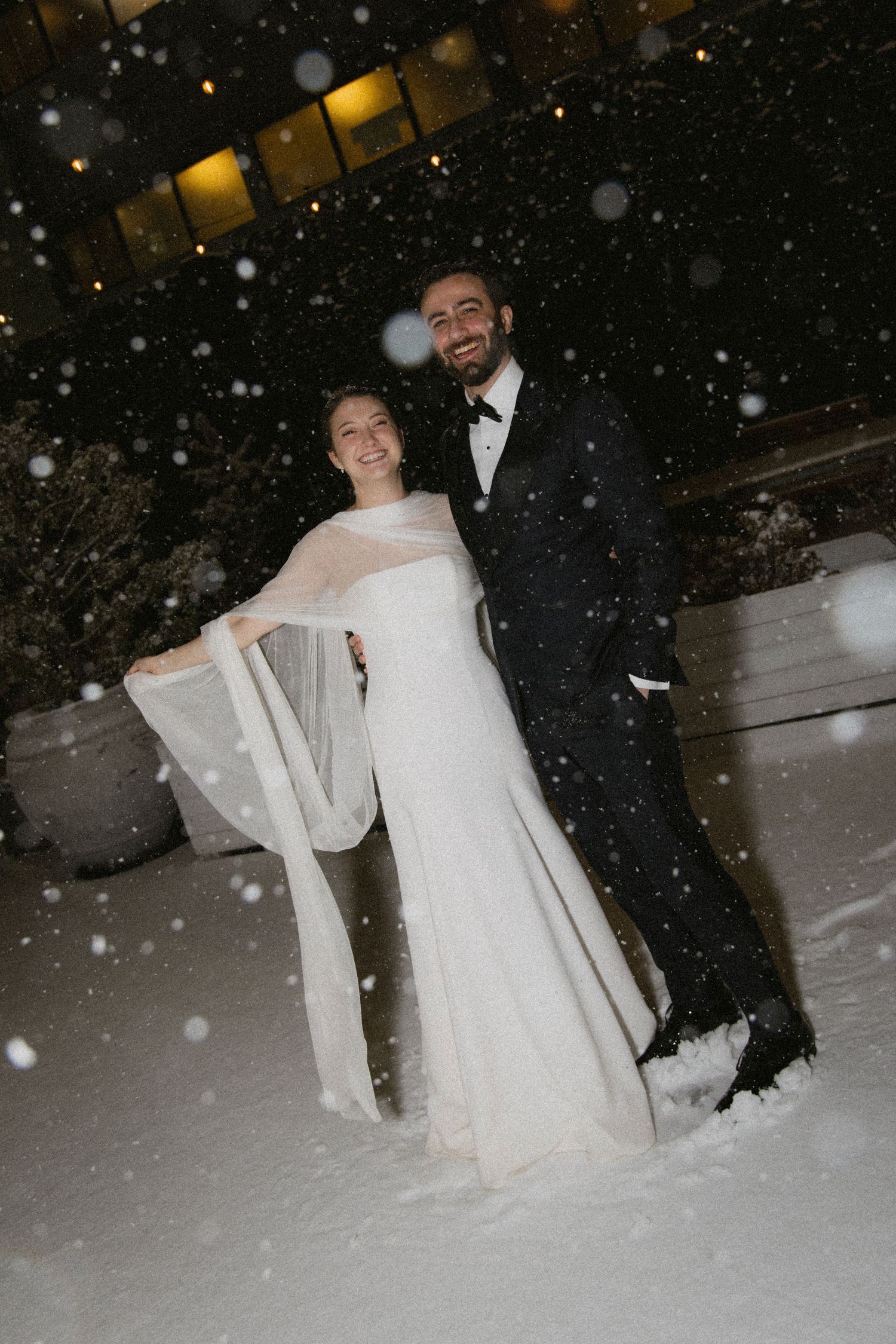 A magical winter wedding photo with falling snow and glowing lights in the background.
