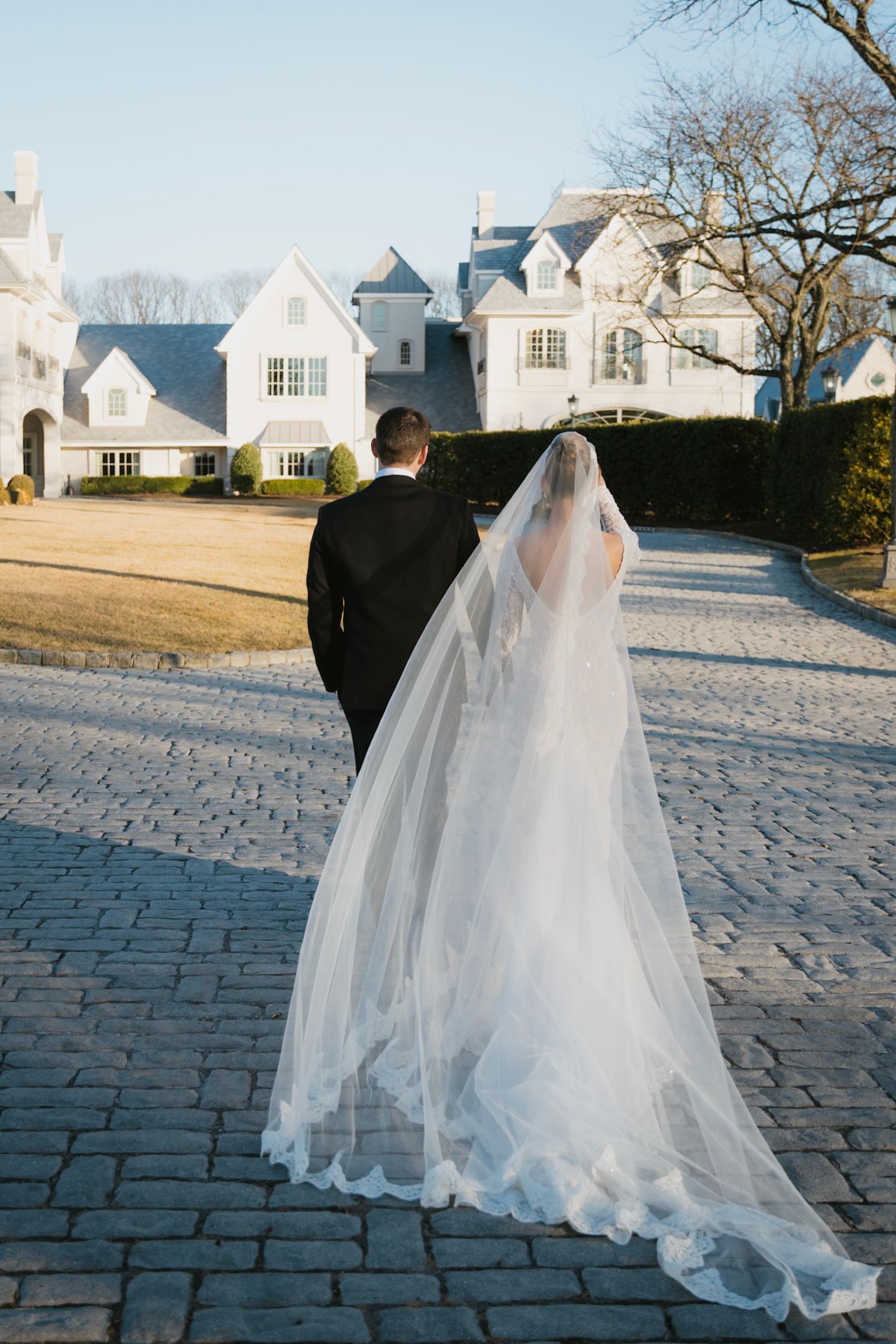 Wedding dress flowing on cobblestone path with white buildings in background on cloudy winter day.