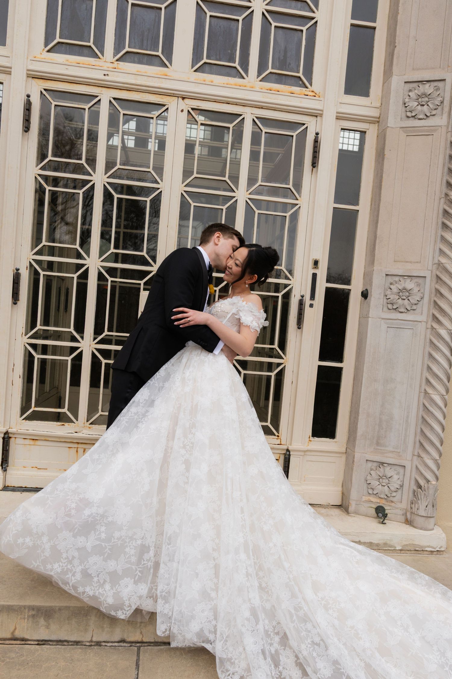 A beautiful wedding couple embraces in front of an ornate building with decorative windows and architecture.
