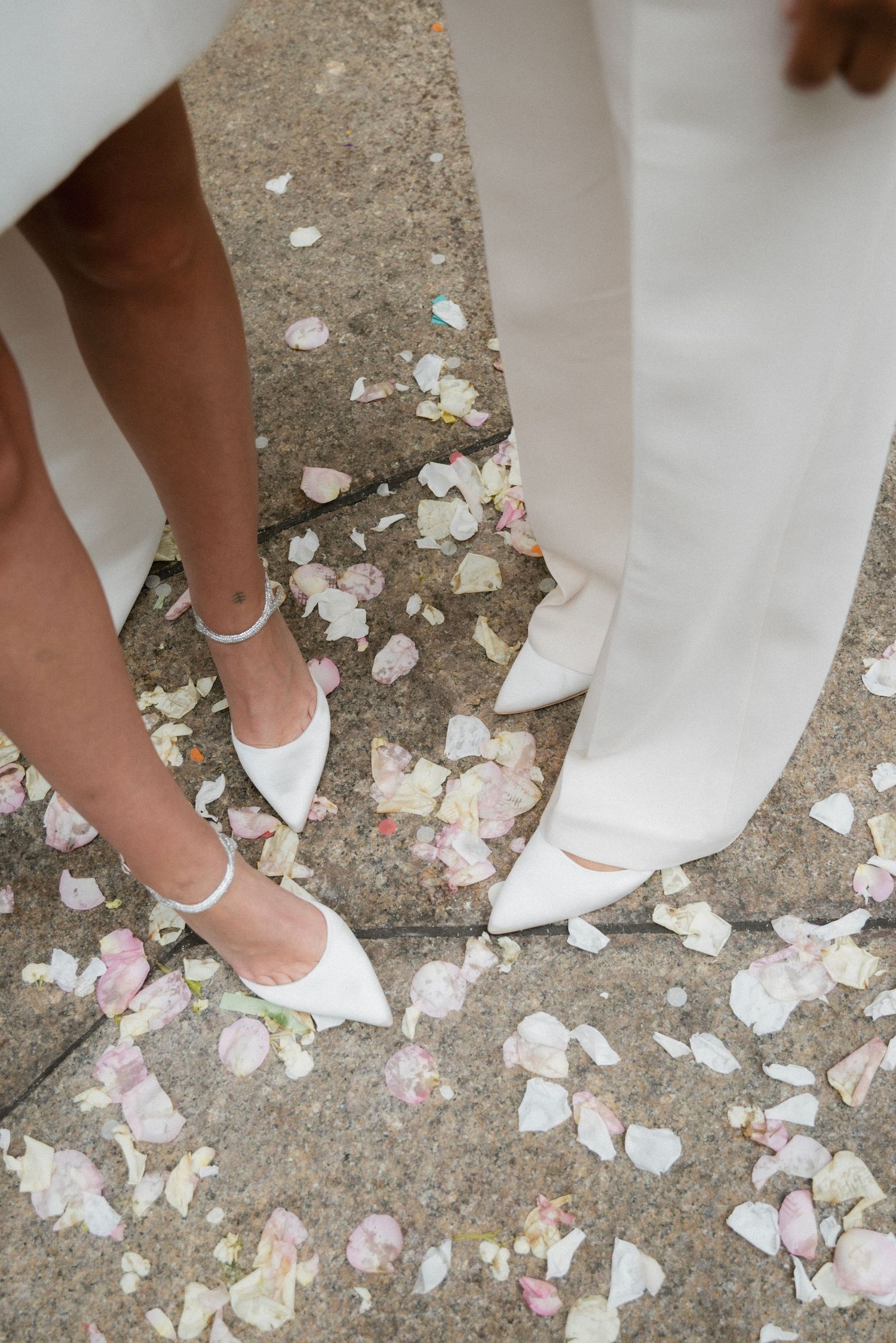 White wedding dress hem and shoes walking on scattered flower petals on concrete ground.