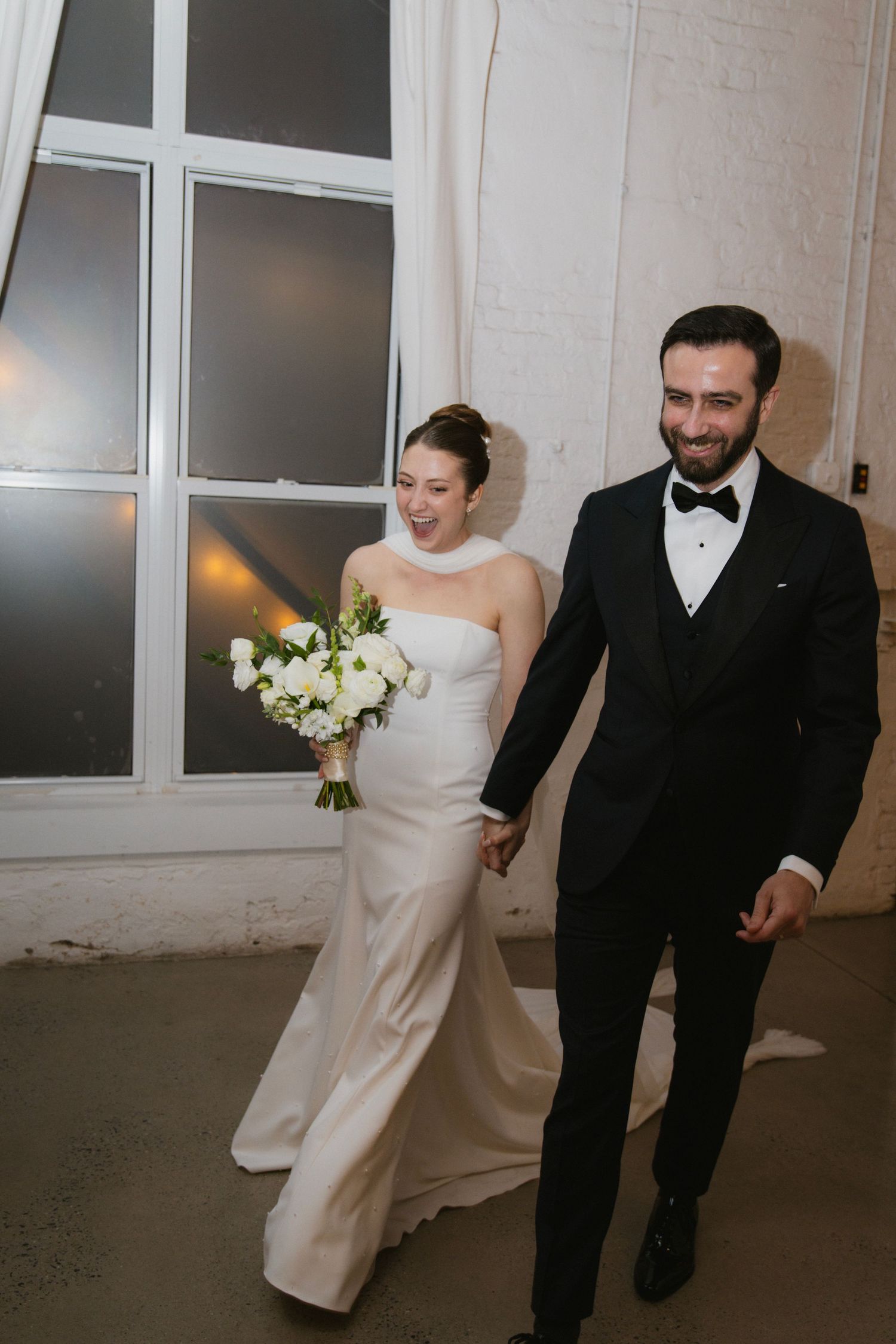 A bride in a white dress and groom in black tuxedo walk together in a modern indoor space.
