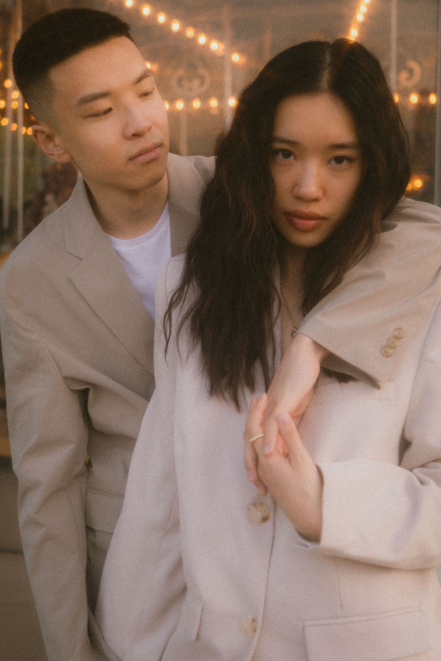 A romantic couple in beige coats pose together against a warm background with string lights.