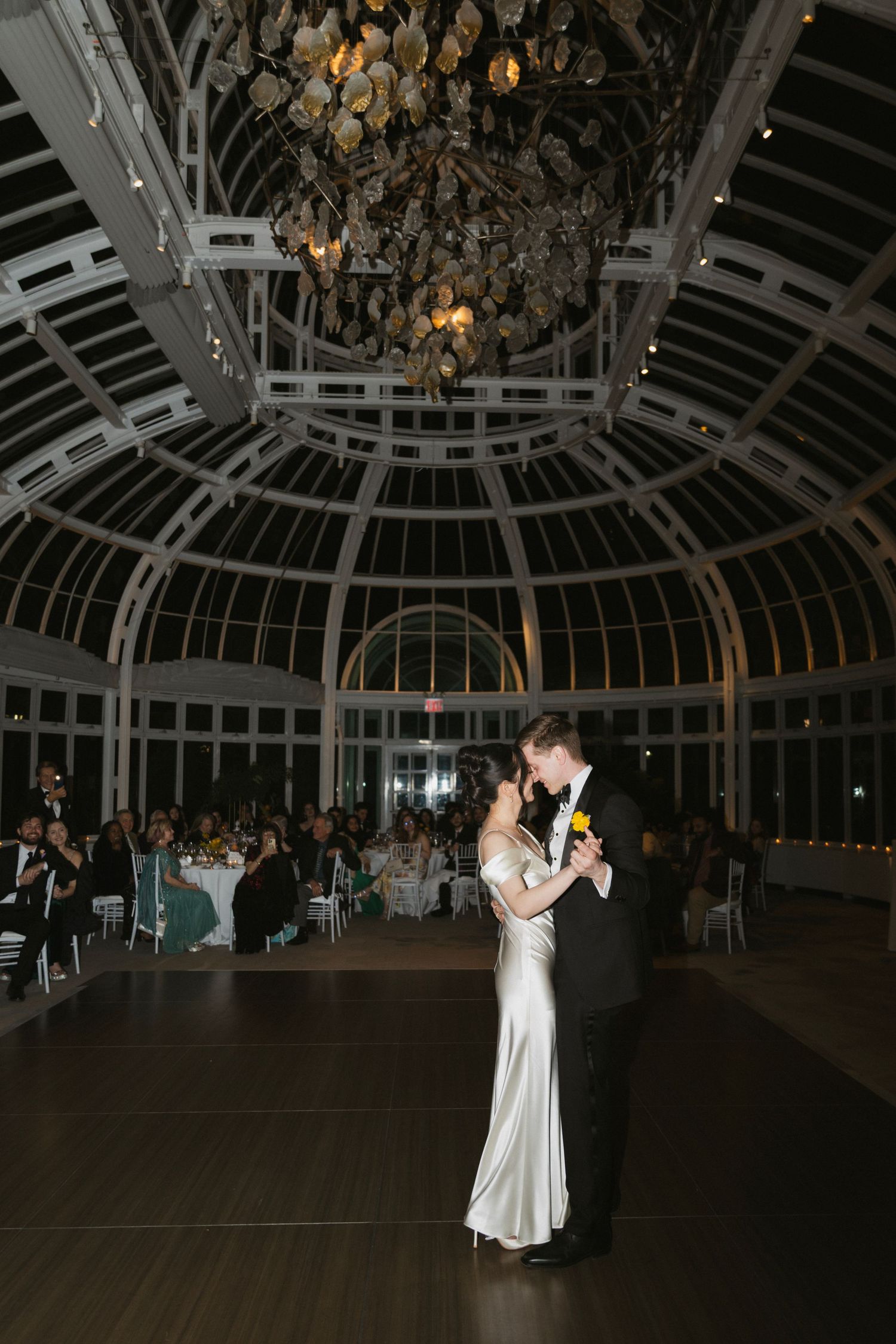Couple shares their first dance under ornate chandelier in a stunning glass-domed conservatory.