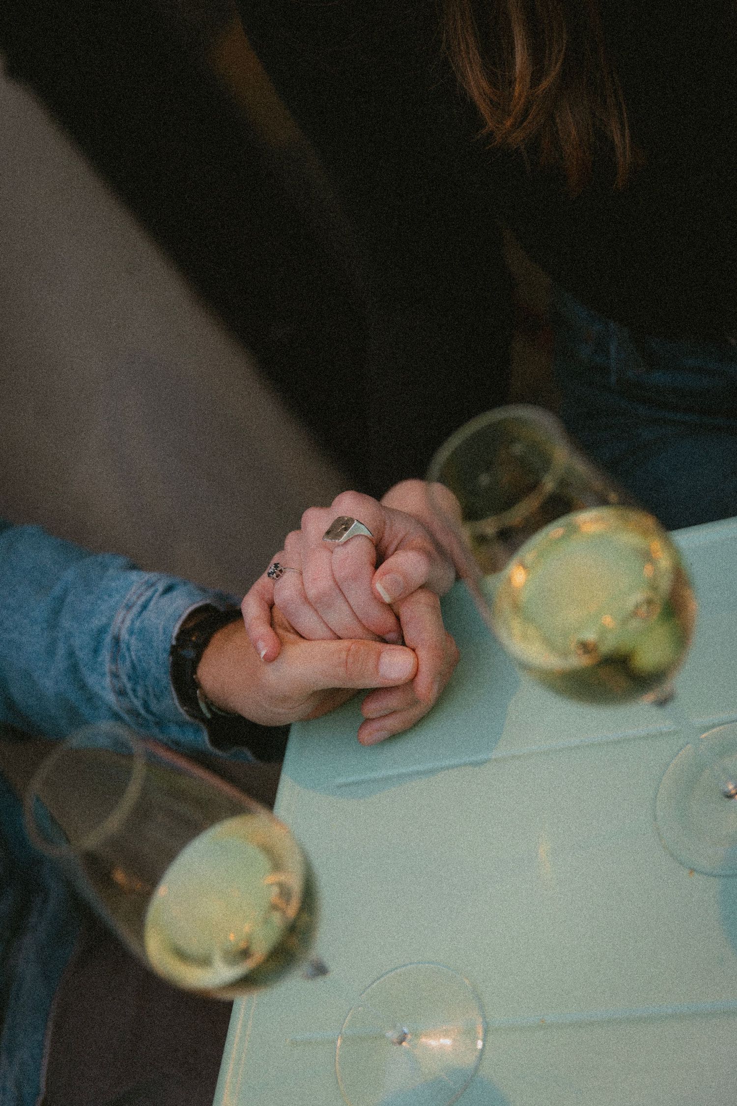 Close-up shot of hands clasped together on a light-colored table with wine glasses in soft lighting.