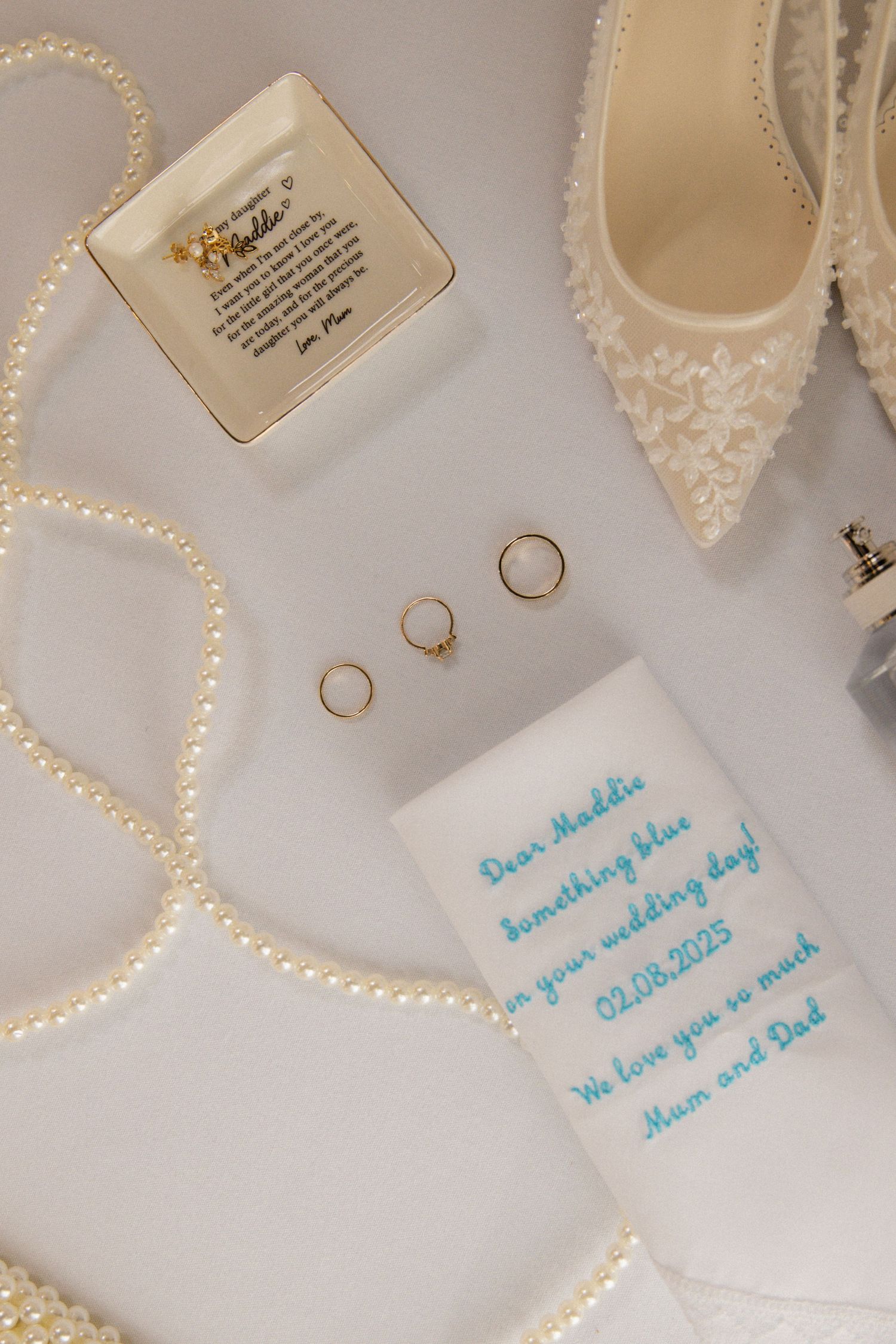 Wedding details including rings, pearl necklace, lace shoes and personalized handkerchief on light background.