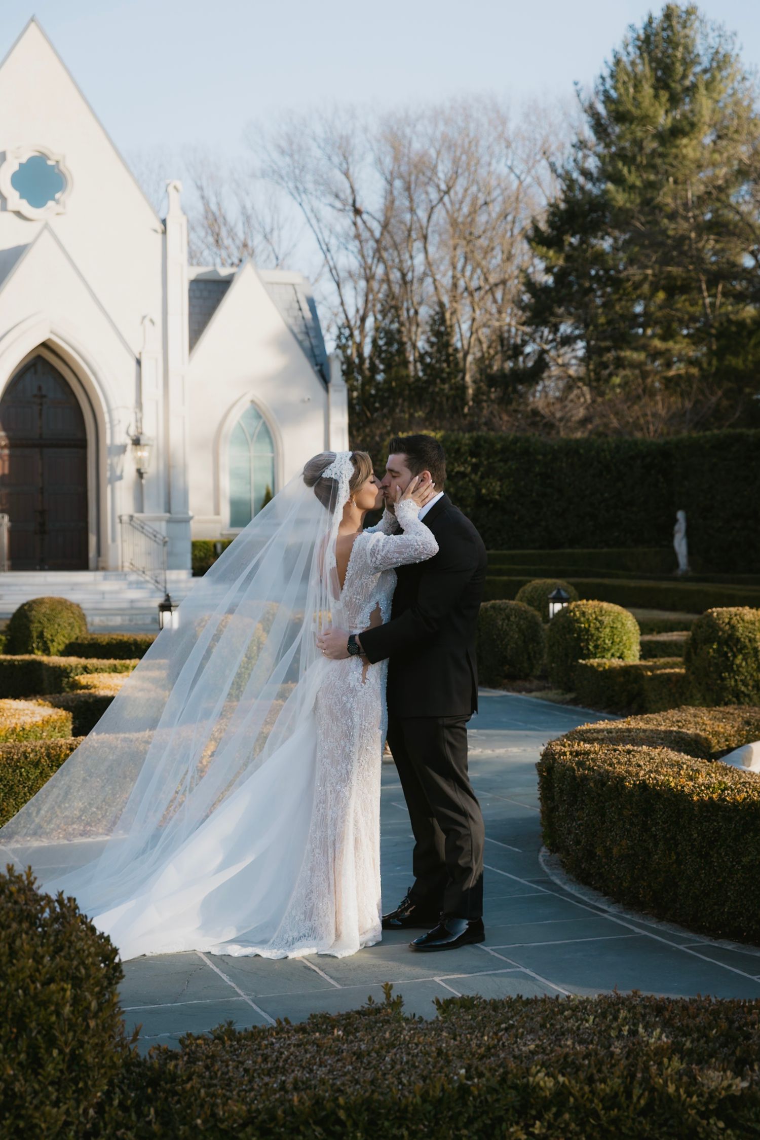A dramatic cathedral veil catches the wind outside a white church on a wedding day.