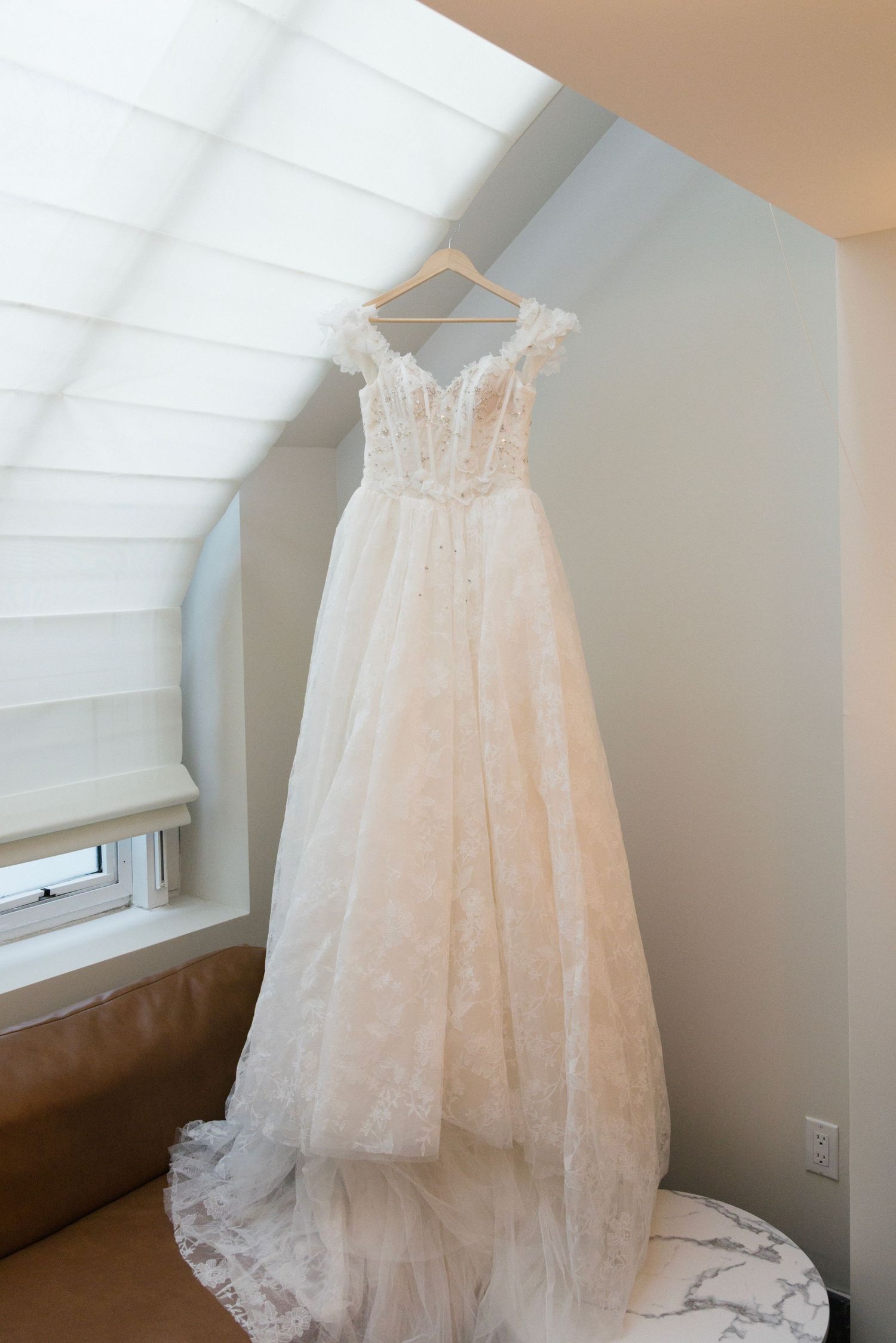 An elegant white lace wedding gown hanging in a sunlit room.
