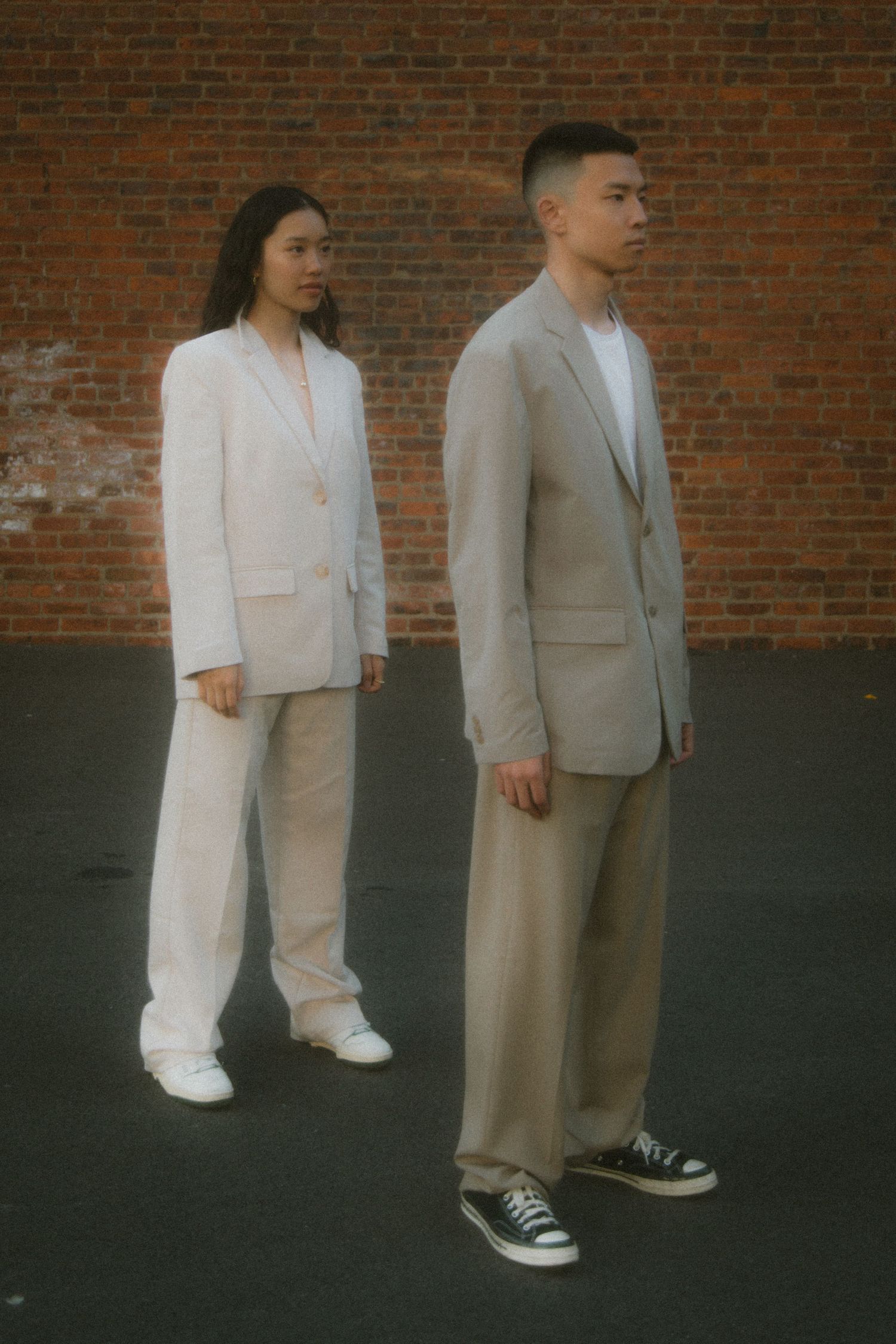 Two people in light colored business suits and casual sneakers pose against a brick wall in moody lighting.