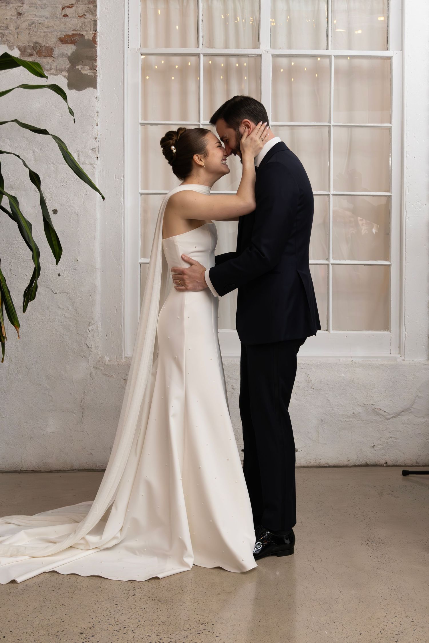 An intimate wedding moment captured as a couple embraces near a white window frame.