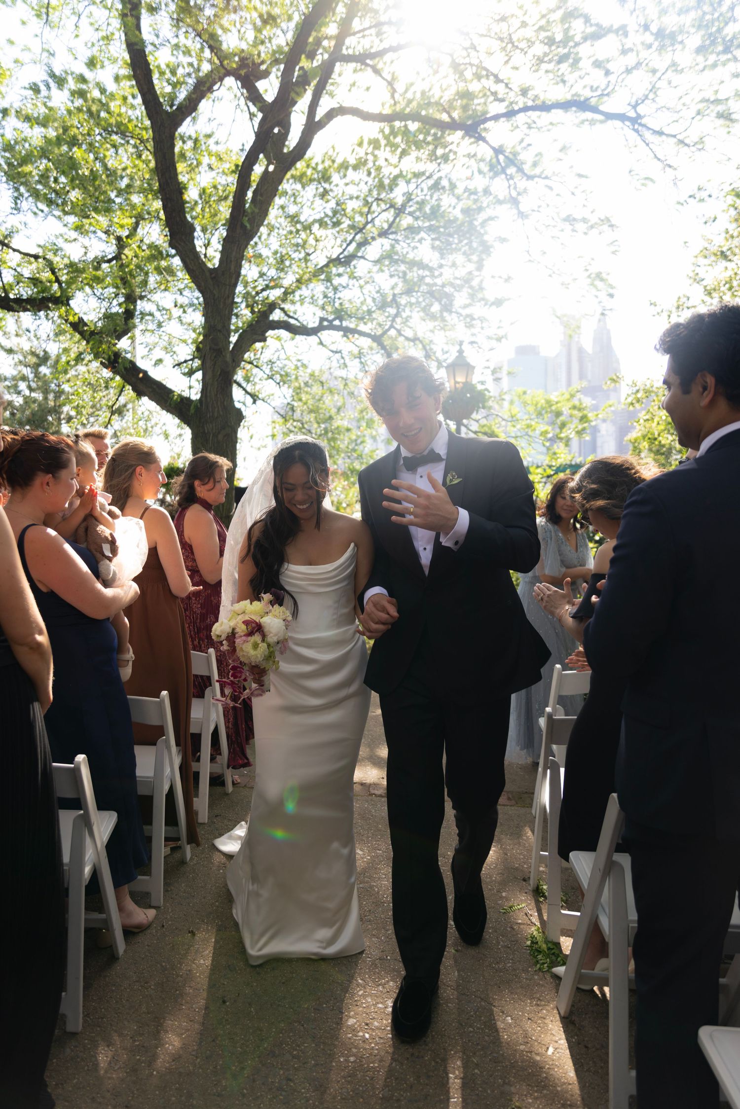 Bride and groom walk down outdoor aisle after wedding ceremony, guests applauding under sunlit trees.