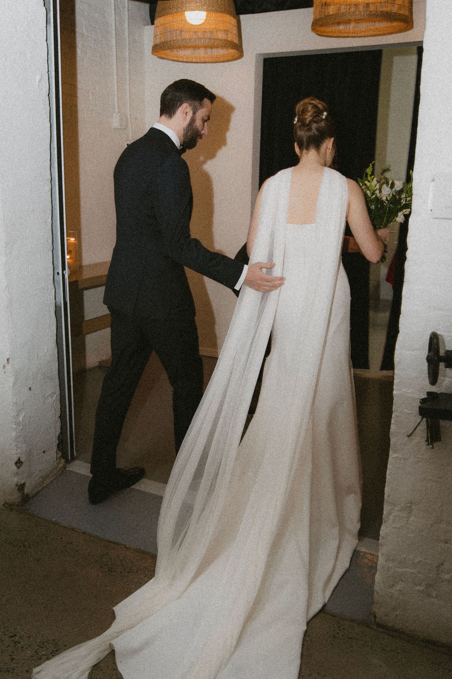 A bride in an elegant white gown with long train walks through doorway guided by groom.