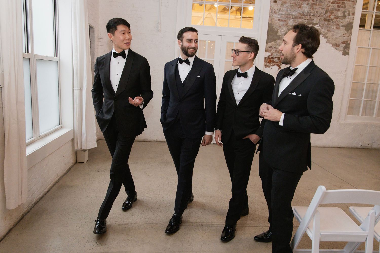 Groomsmen in black tuxedos walking and laughing together in an elegant indoor venue.