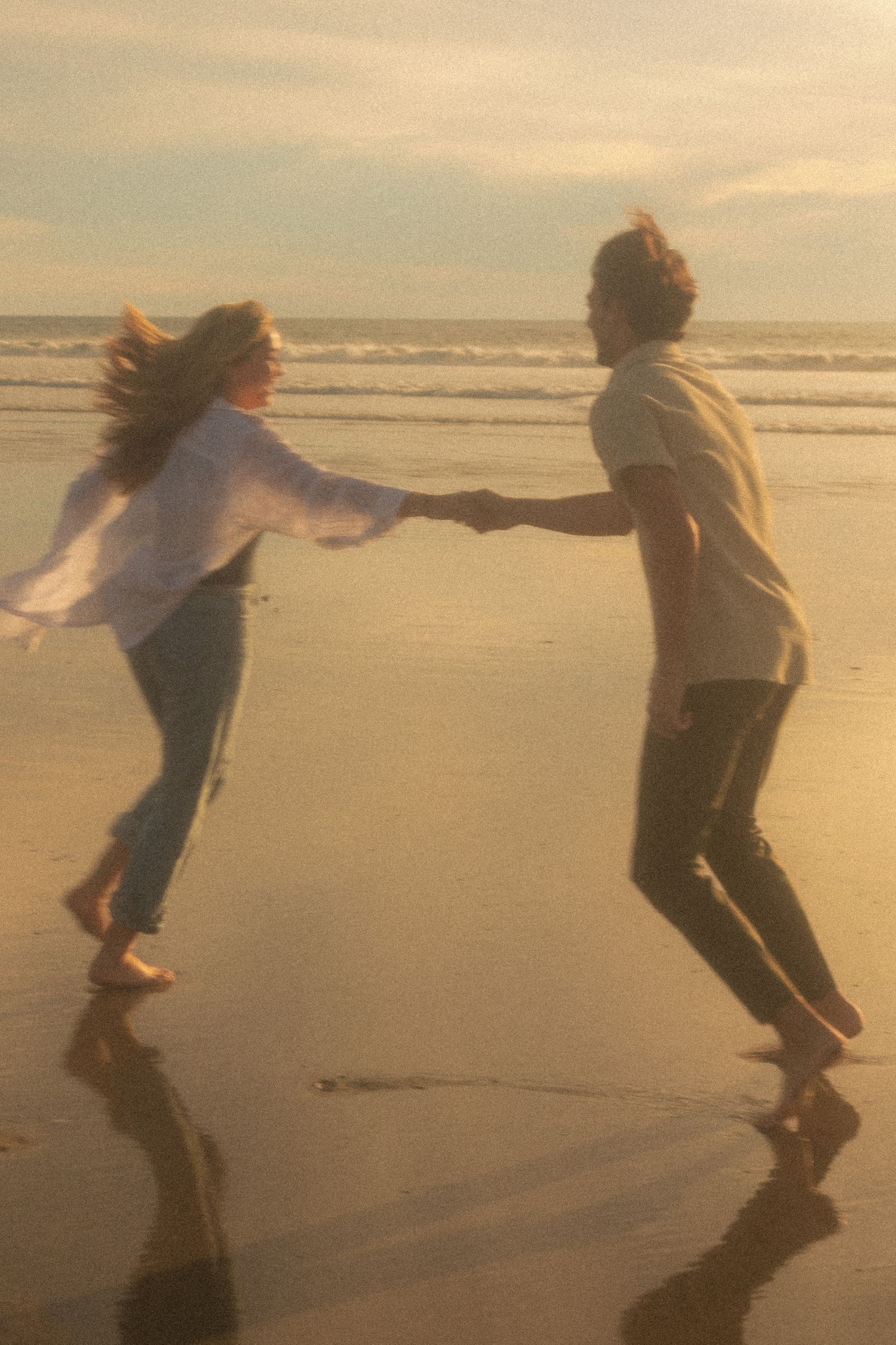 Couple playfully dancing on a beach at sunset with waves in the background.