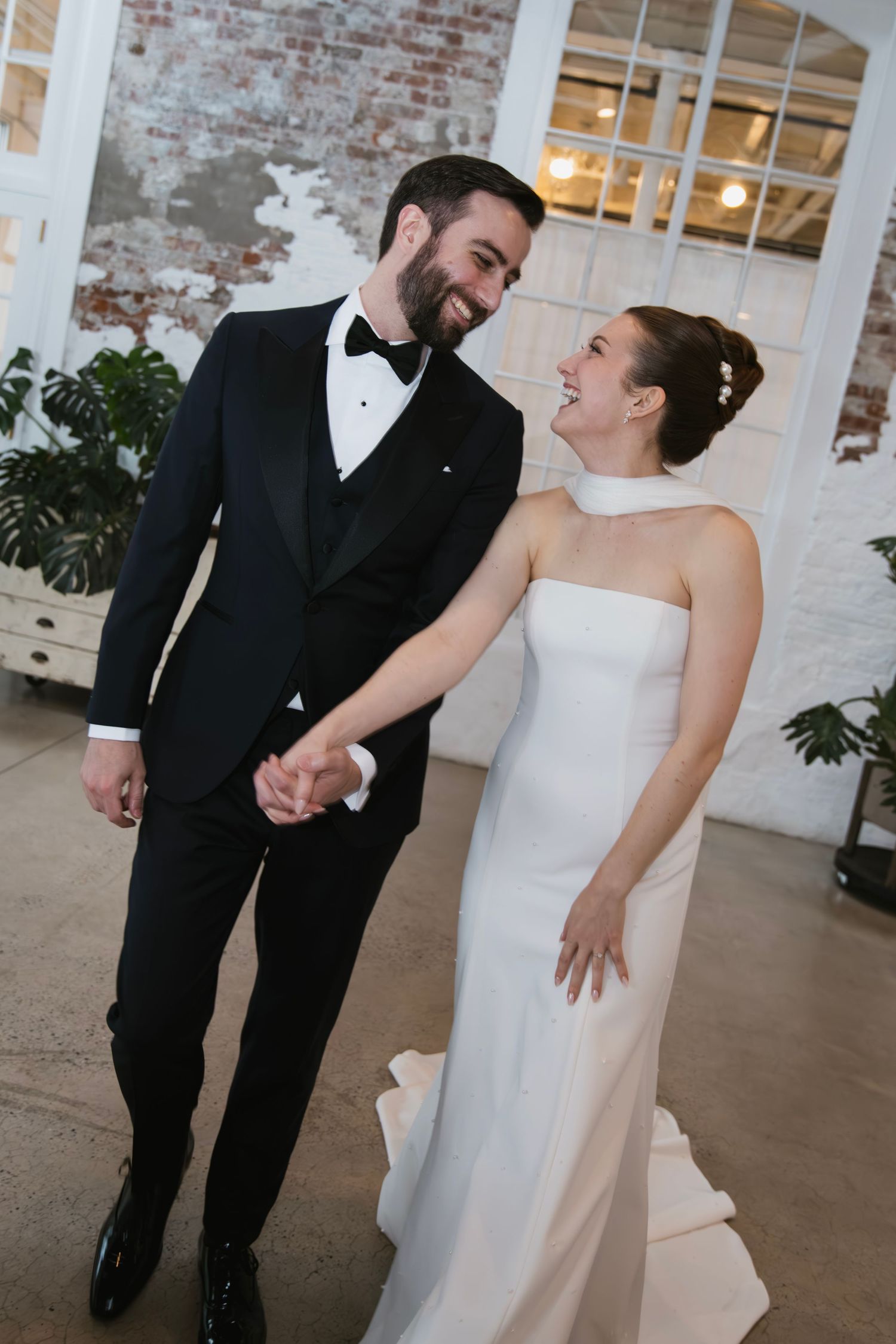 A joyful couple sharing a laugh in their wedding attire against an industrial brick wall backdrop.