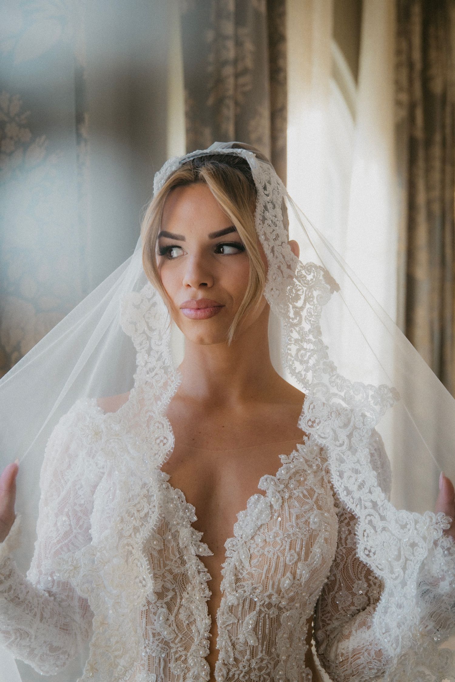 A bride wearing an elegant white lace wedding dress with a plunging neckline and delicate veil in soft window light.