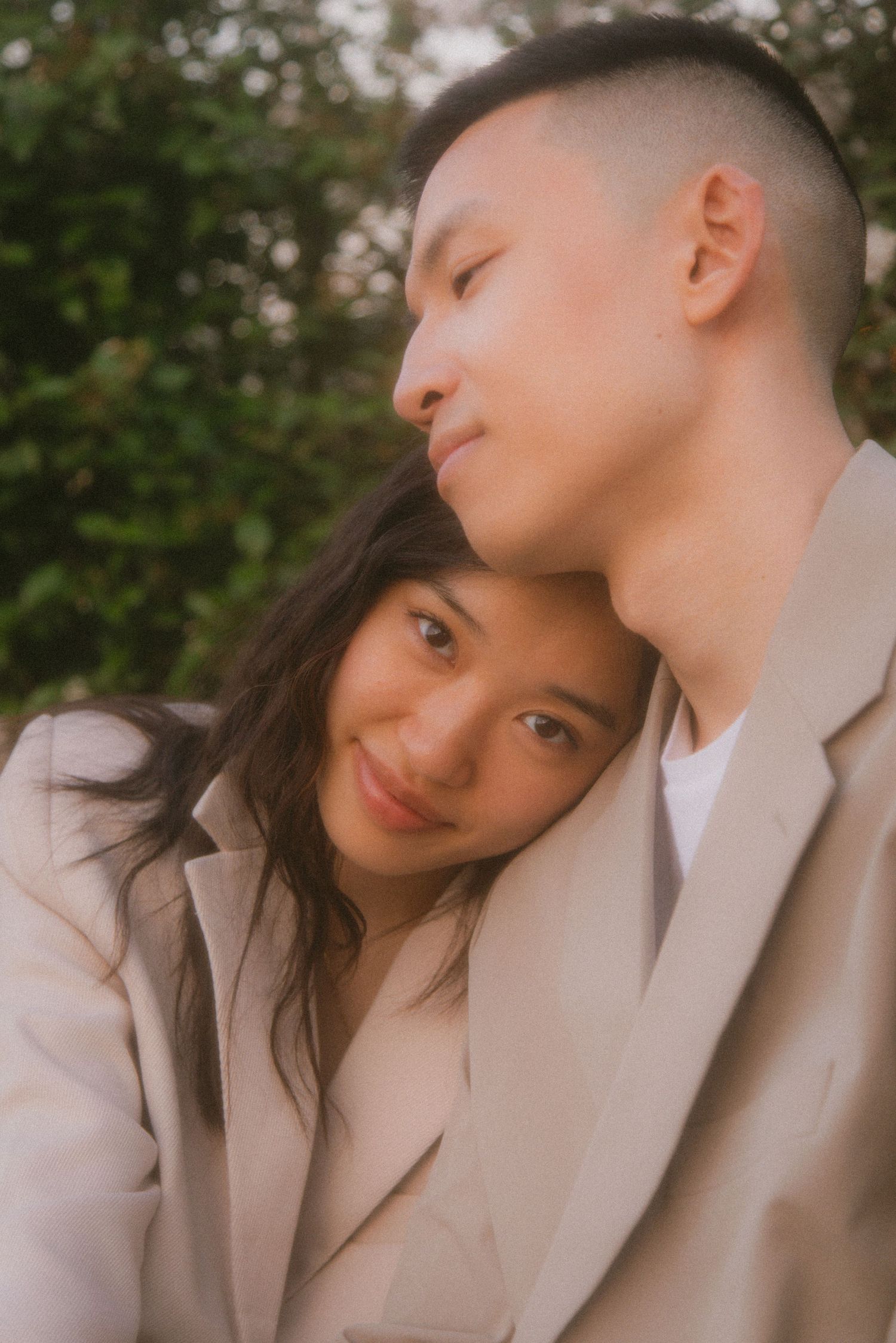 A couple in matching beige coats share an intimate moment while embracing outdoors with trees in the background.
