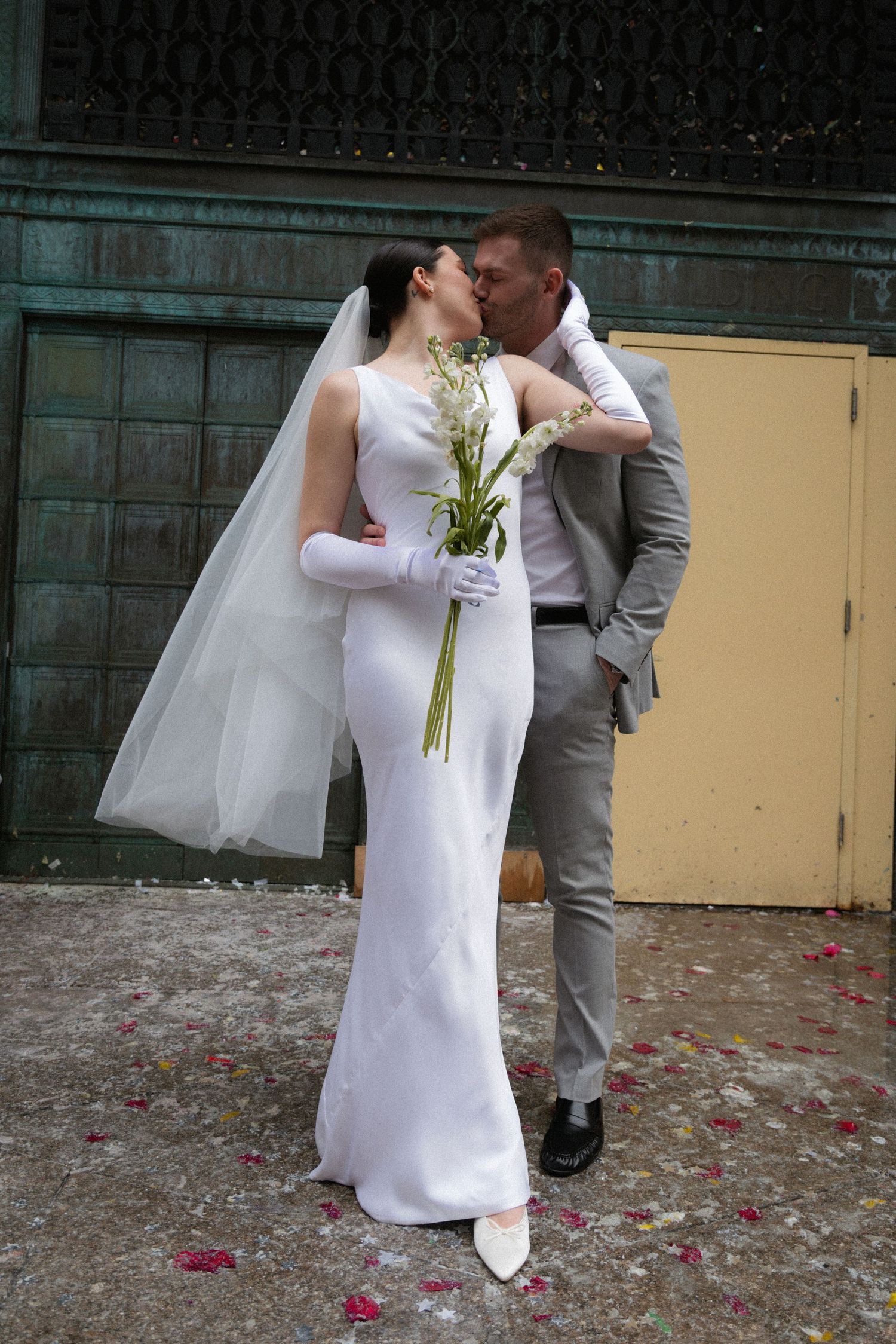 Bride in white gown and veil kisses groom in gray suit outdoors, surrounded by scattered rose petals.