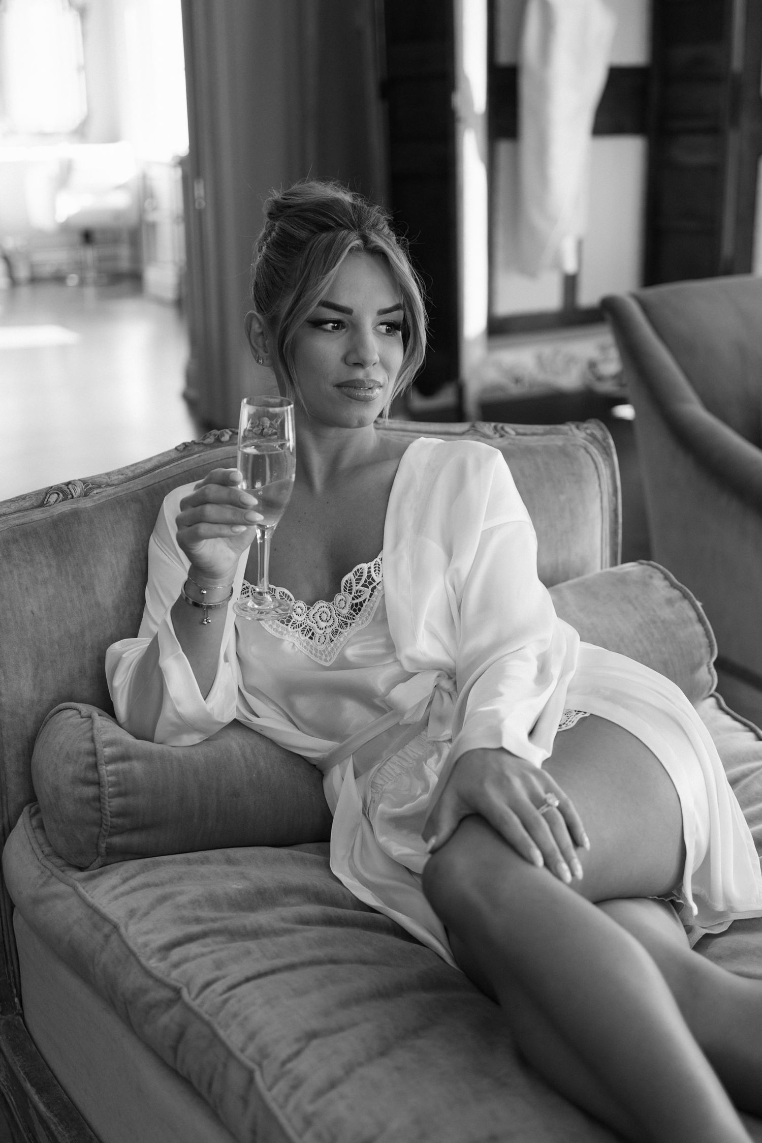 Woman in white silk robe relaxing on couch with champagne glass in elegant setting.