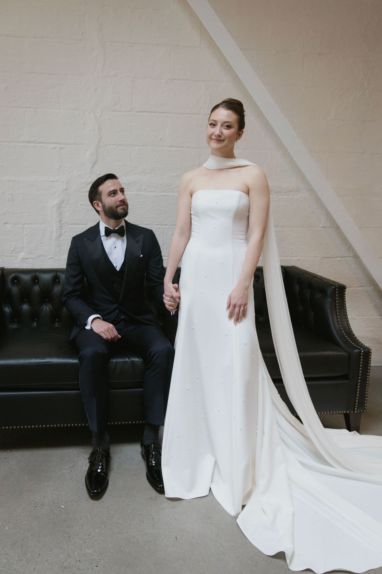 A bride in a strapless white gown with a flowing train stands beside a groom in black tuxedo seated on a leather couch.